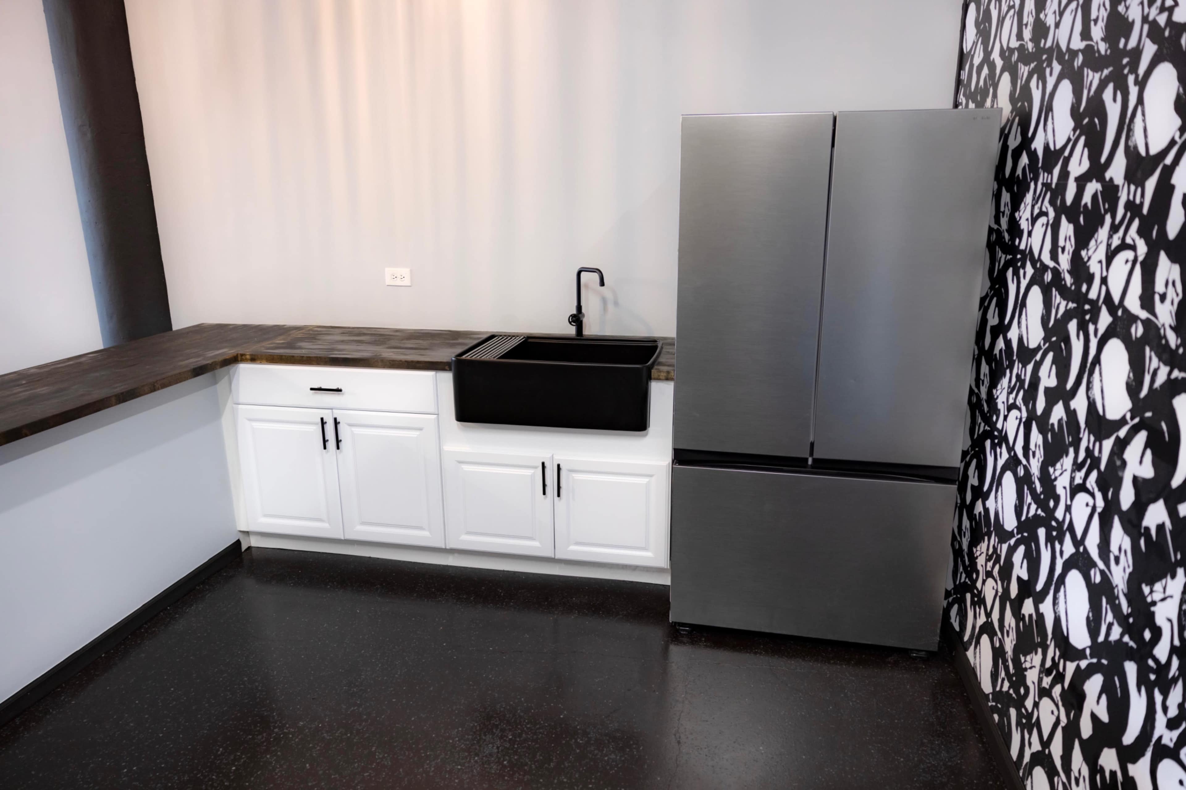The image shows a modern kitchen with a black sink and countertop, white cabinets, and a stainless steel refrigerator against a patterned wall.