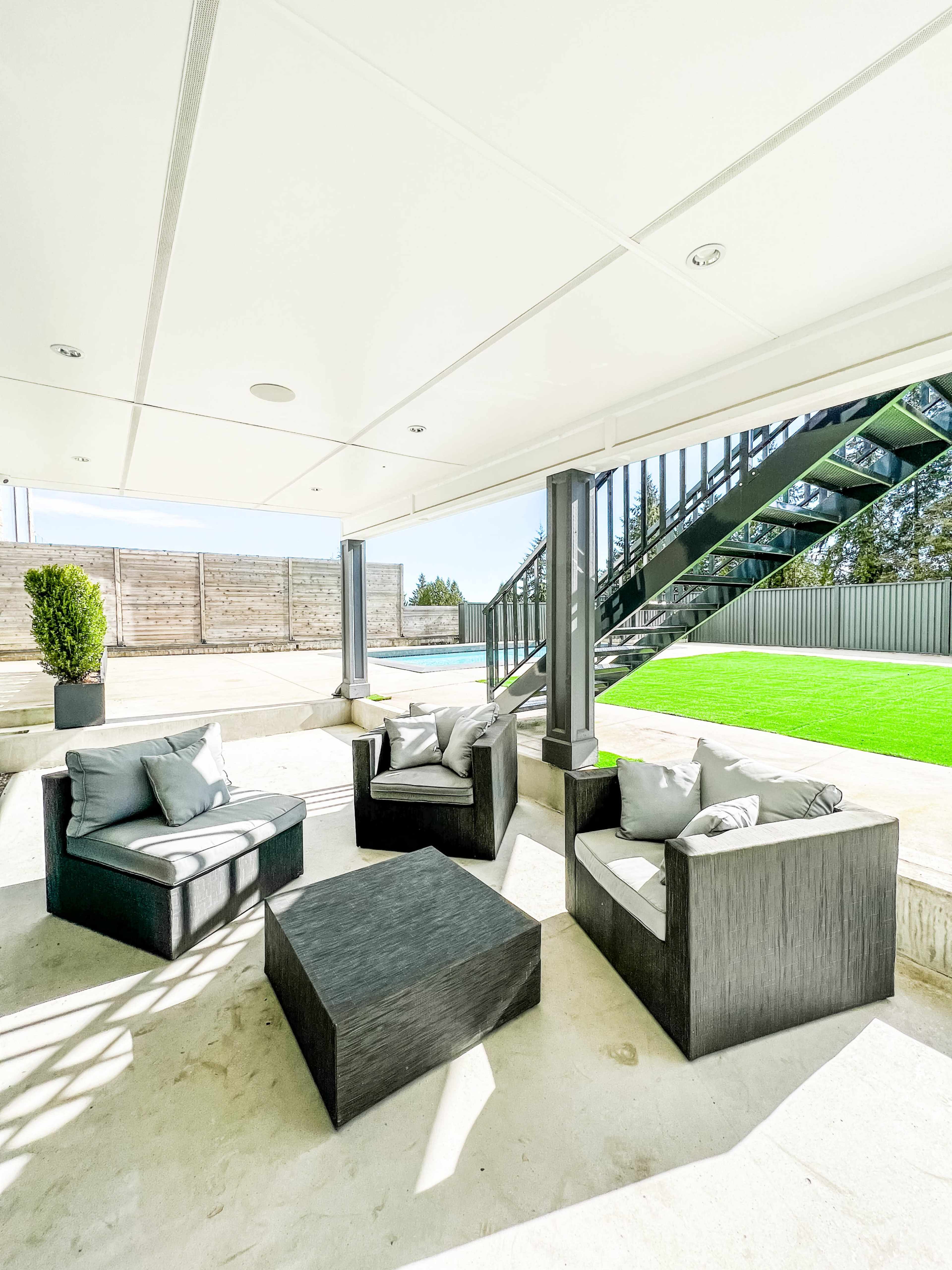 A modern outdoor seating area with black furniture arranged around a square table, set beneath a covered patio near a pool area.