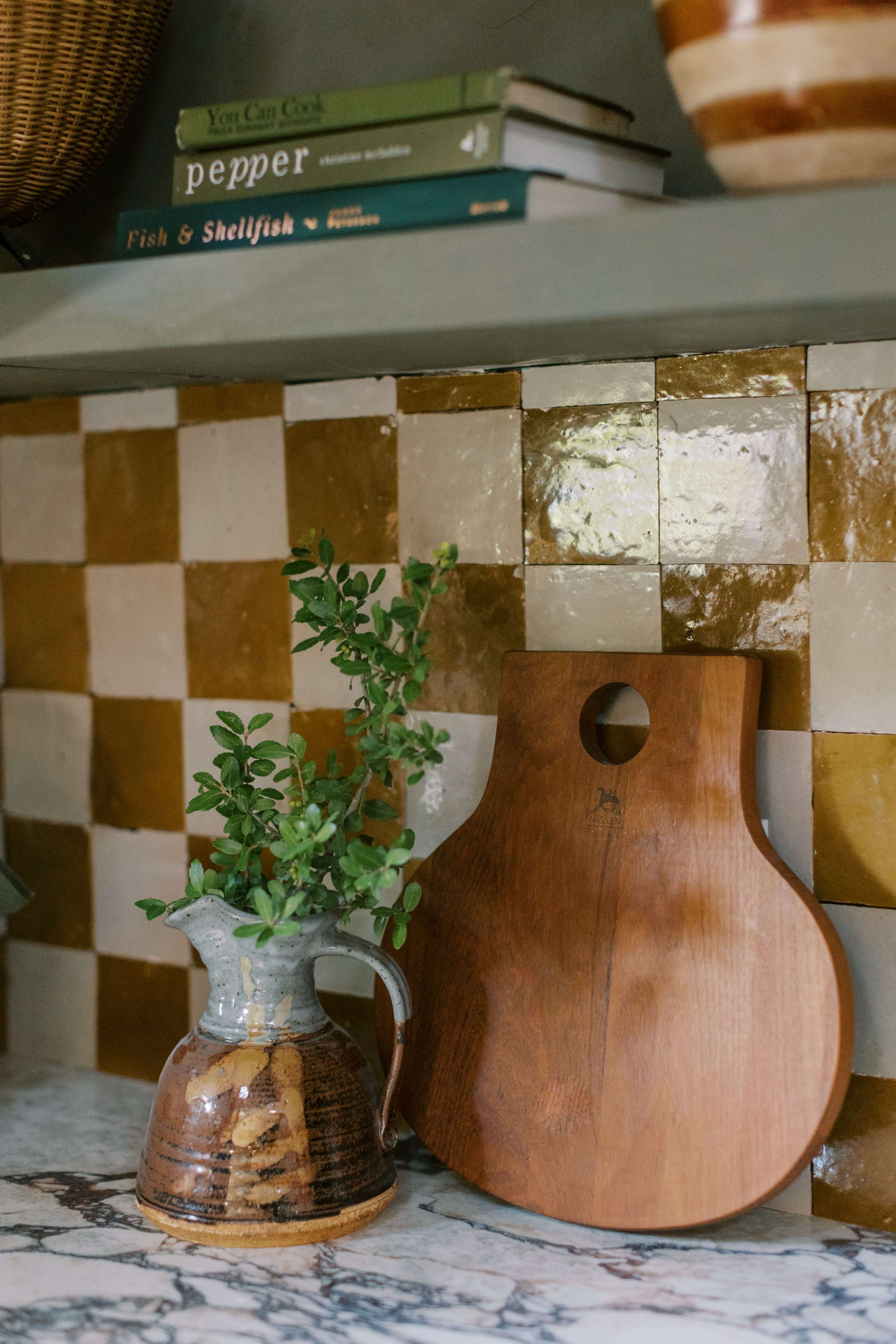 A patterned backsplash in a kitchen displays a ceramic pitcher with a plant and a wooden cutting board on a marble countertop.