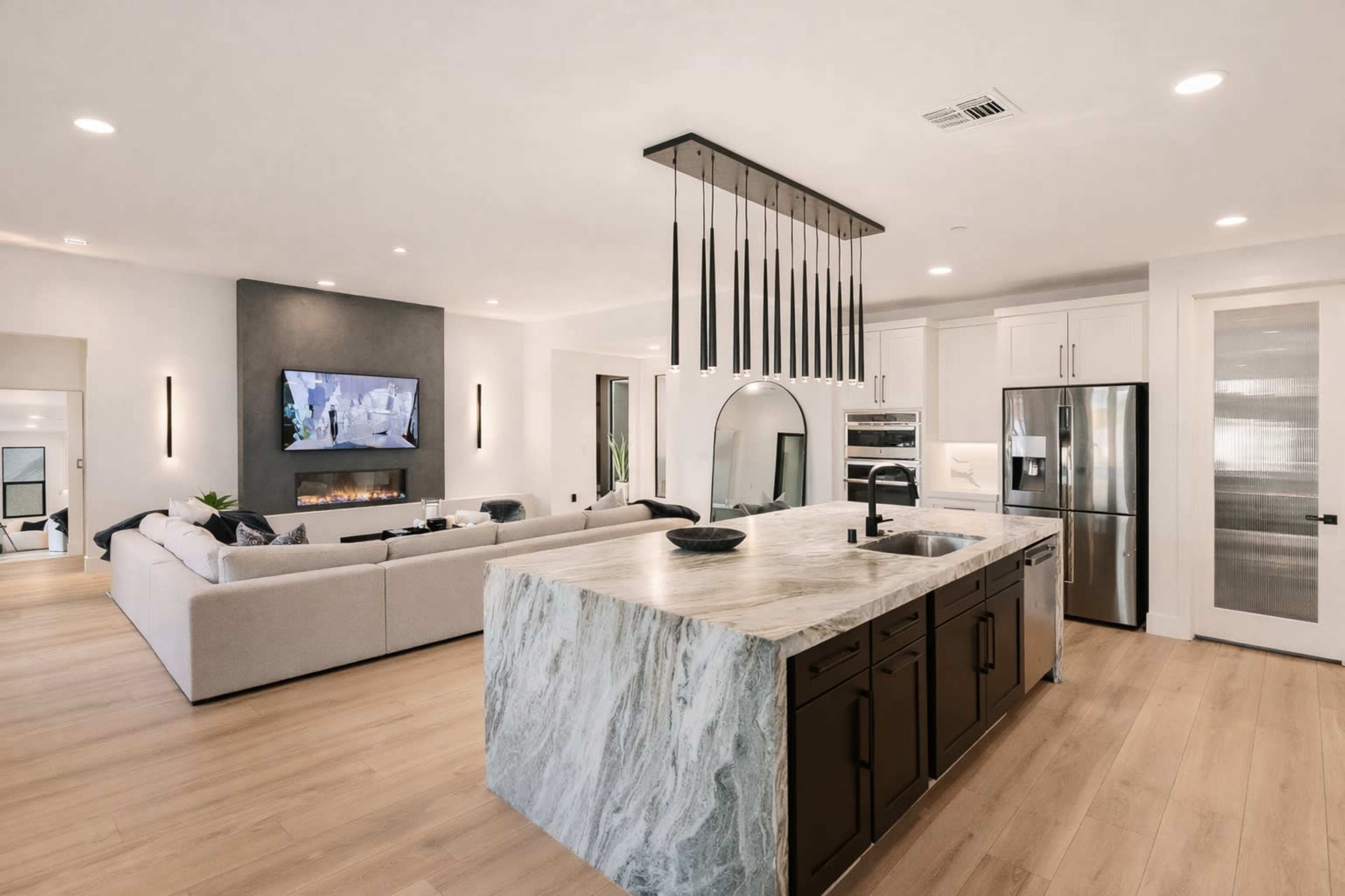 The image shows a modern, open-concept living space with a kitchen island, a living area featuring a large couch, and a wall-mounted television.