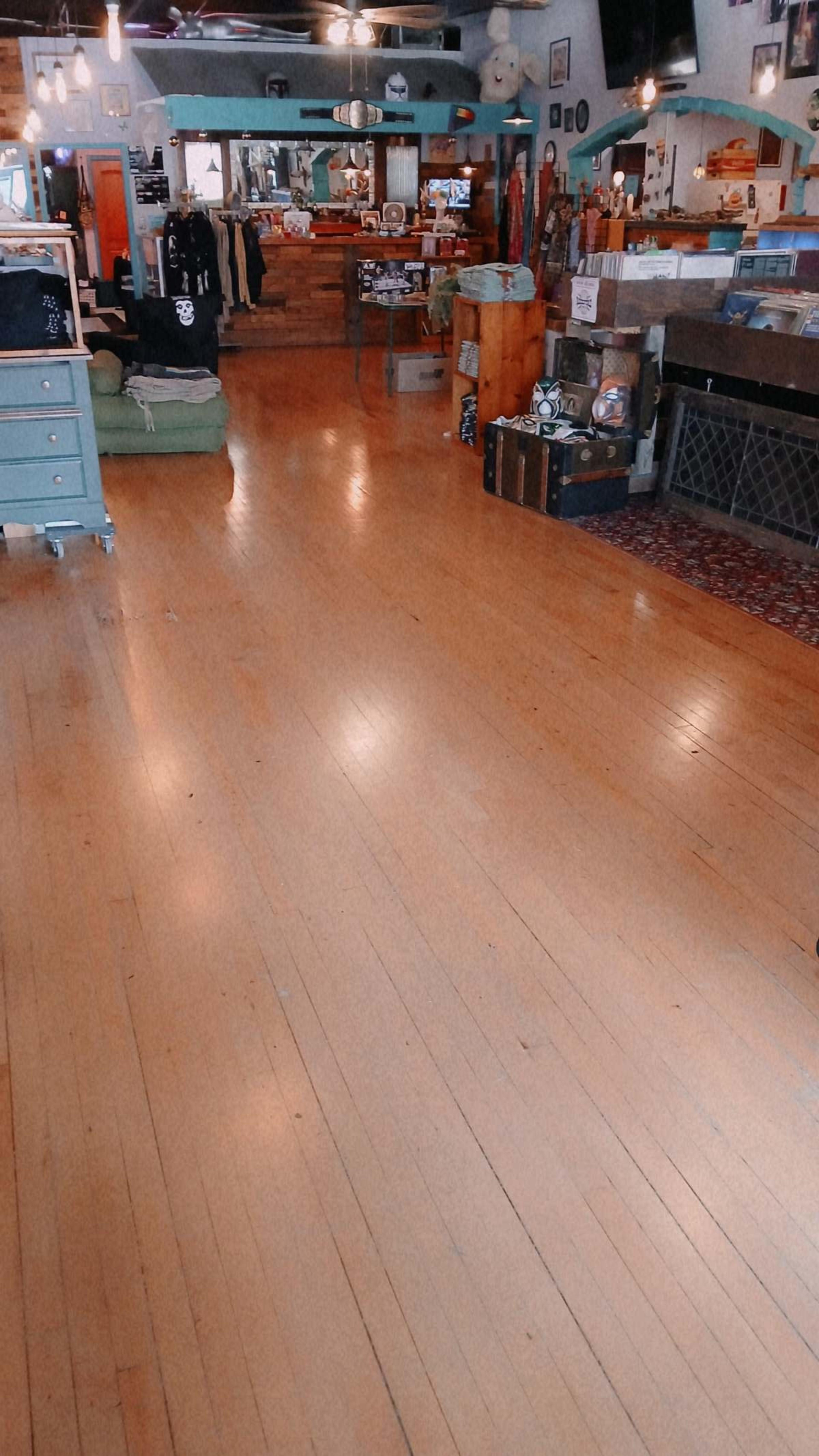The image shows an interior of a retail store with wooden floors and various items, such as clothing and decorative pieces, displayed on shelves and tables.