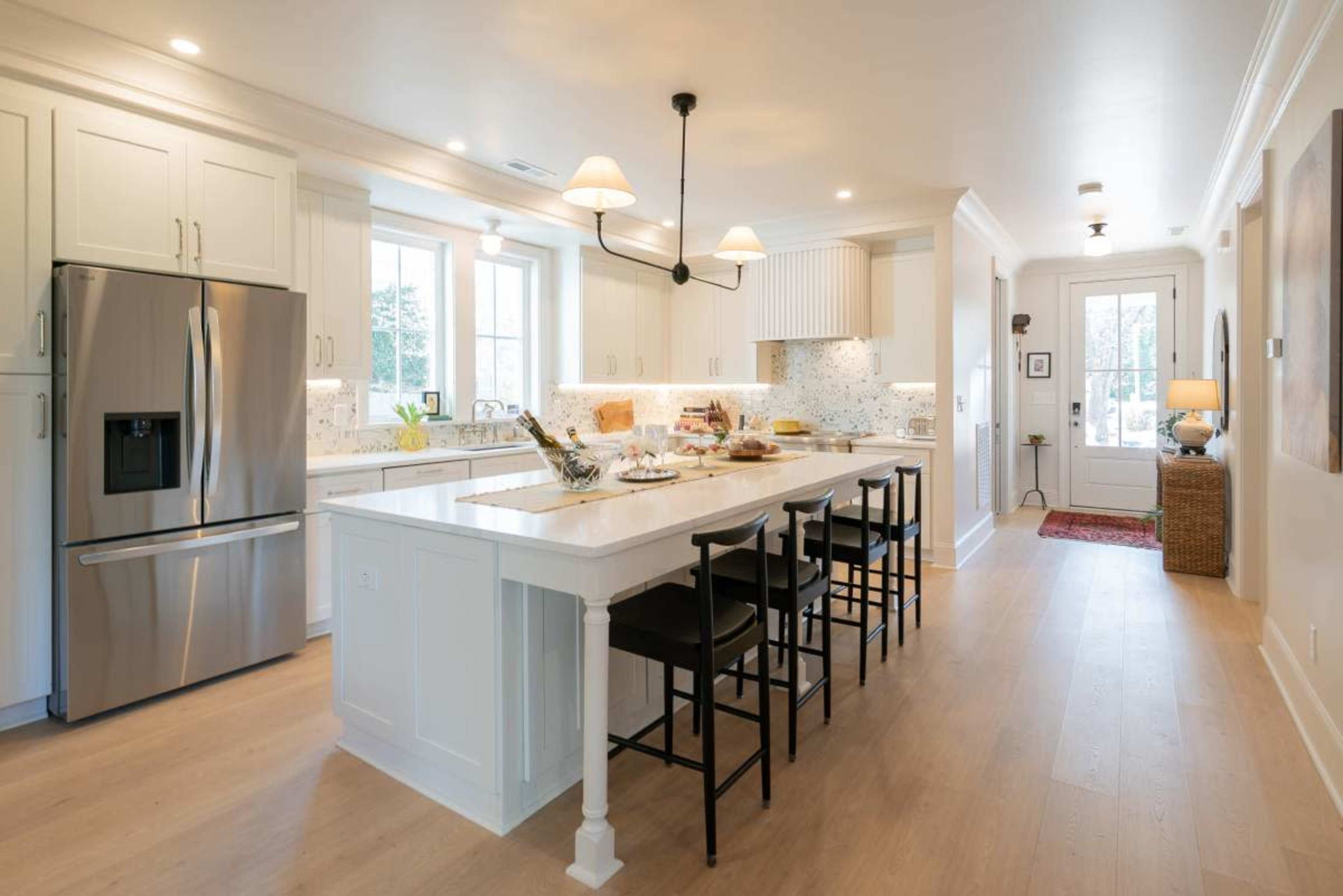 The image shows a modern kitchen with white cabinets, a large island with seating, marble countertops, and stainless steel appliances.