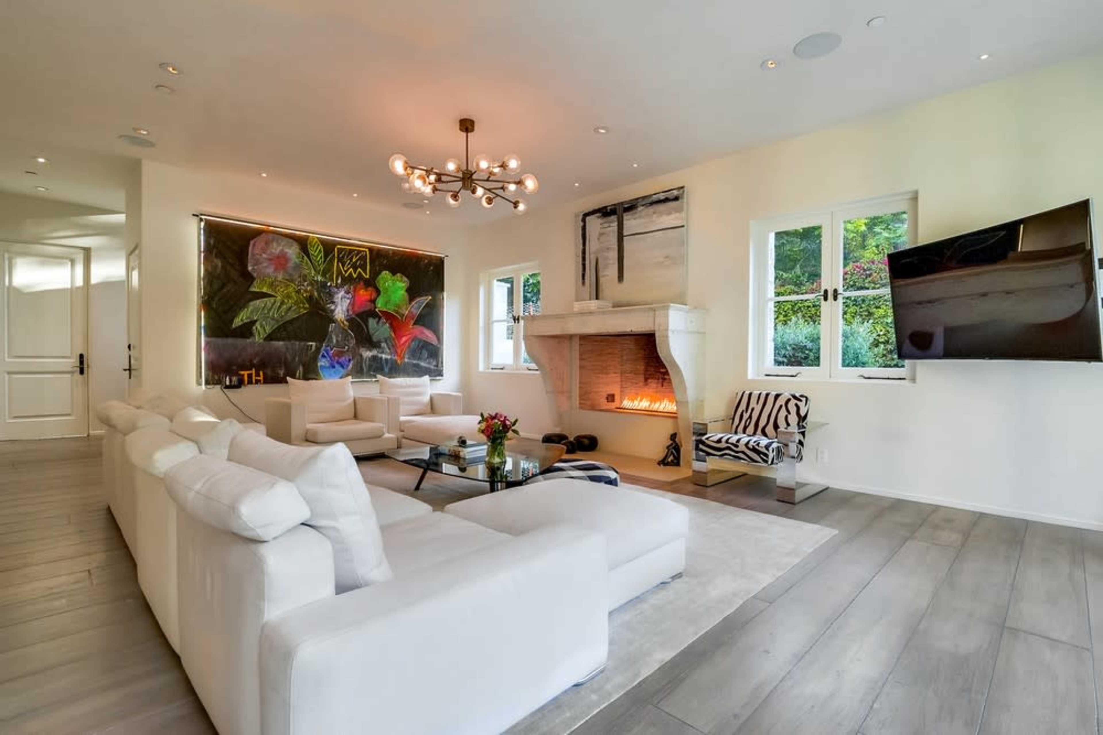 The living room features a white sectional sofa, a modern coffee table, a fireplace surrounded by art, and large windows illuminating the space.