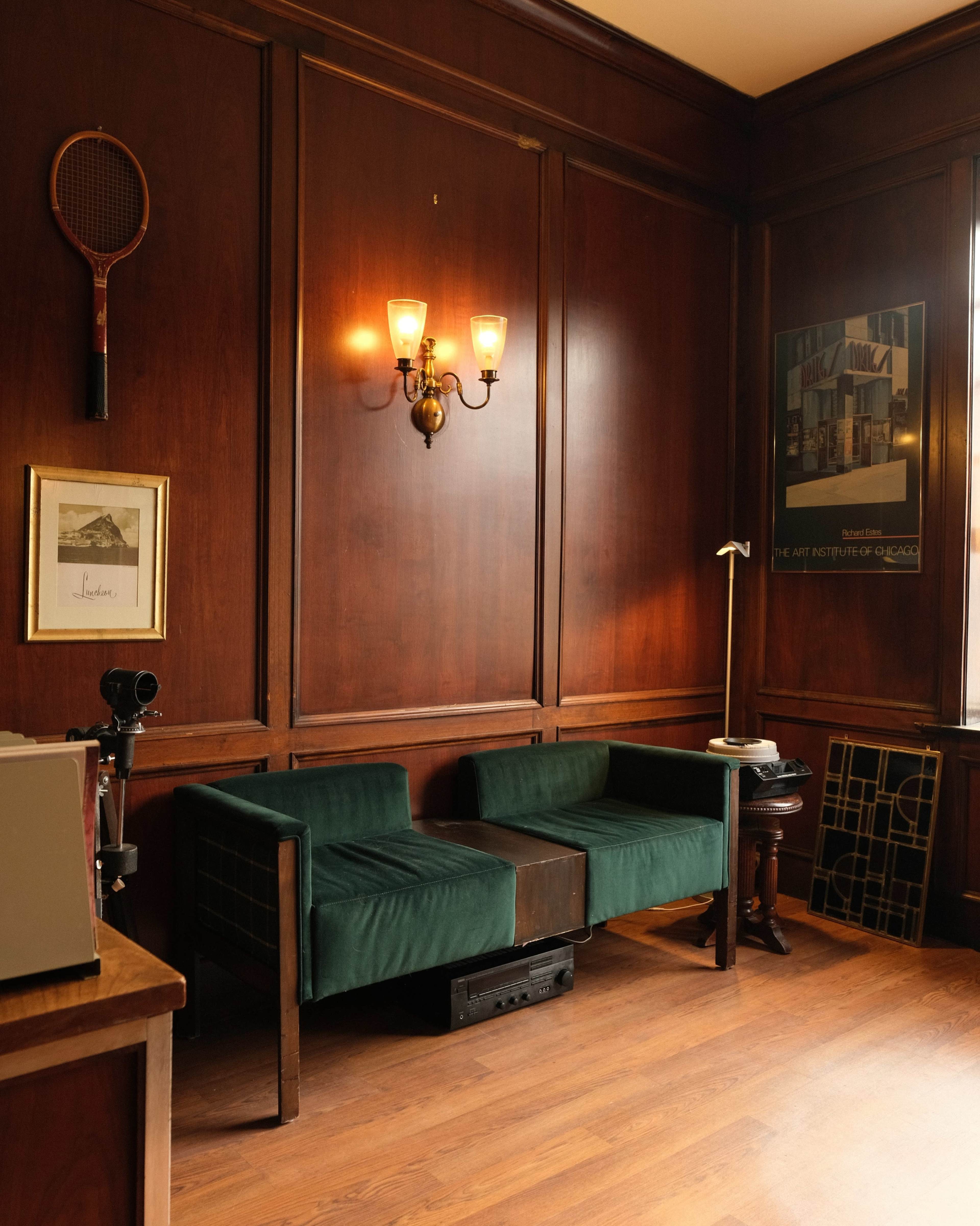 The image shows a wooden-paneled room with two green chairs, a lamp on a small table, a wall-mounted tennis racket, and framed art on the walls.