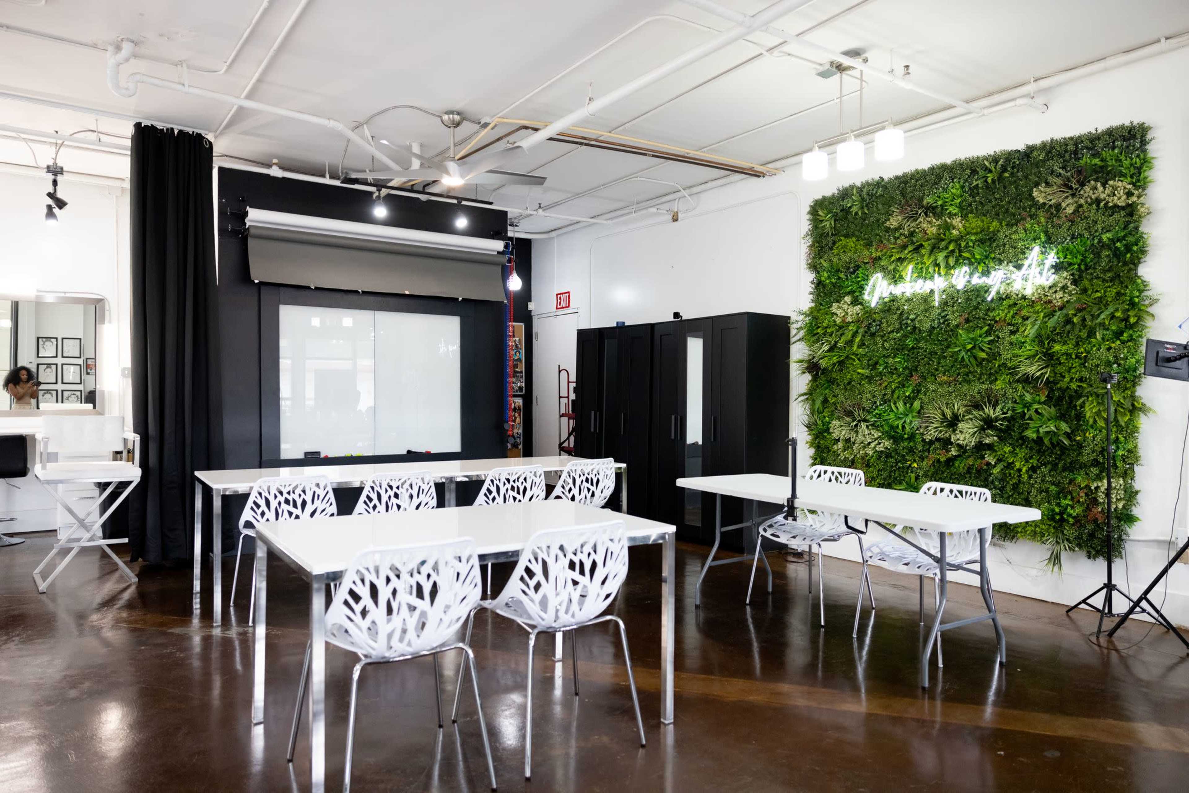 The image shows a modern workspace featuring white tables and chairs, a green vertical garden with a neon sign, and a projector screen on a black wall.