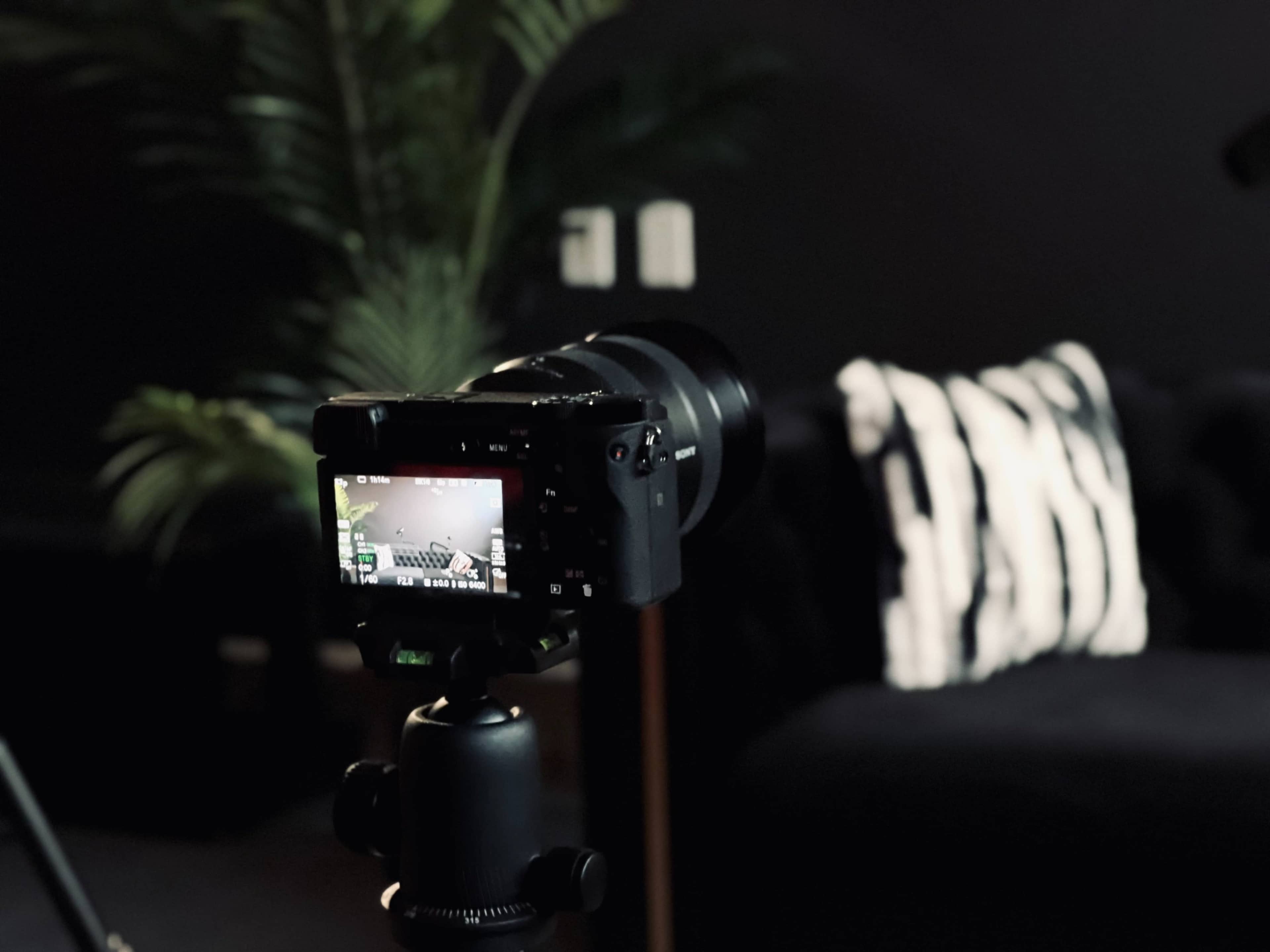 A camera on a tripod is focused on a dark couch with a patterned throw pillow in a dimly lit room.