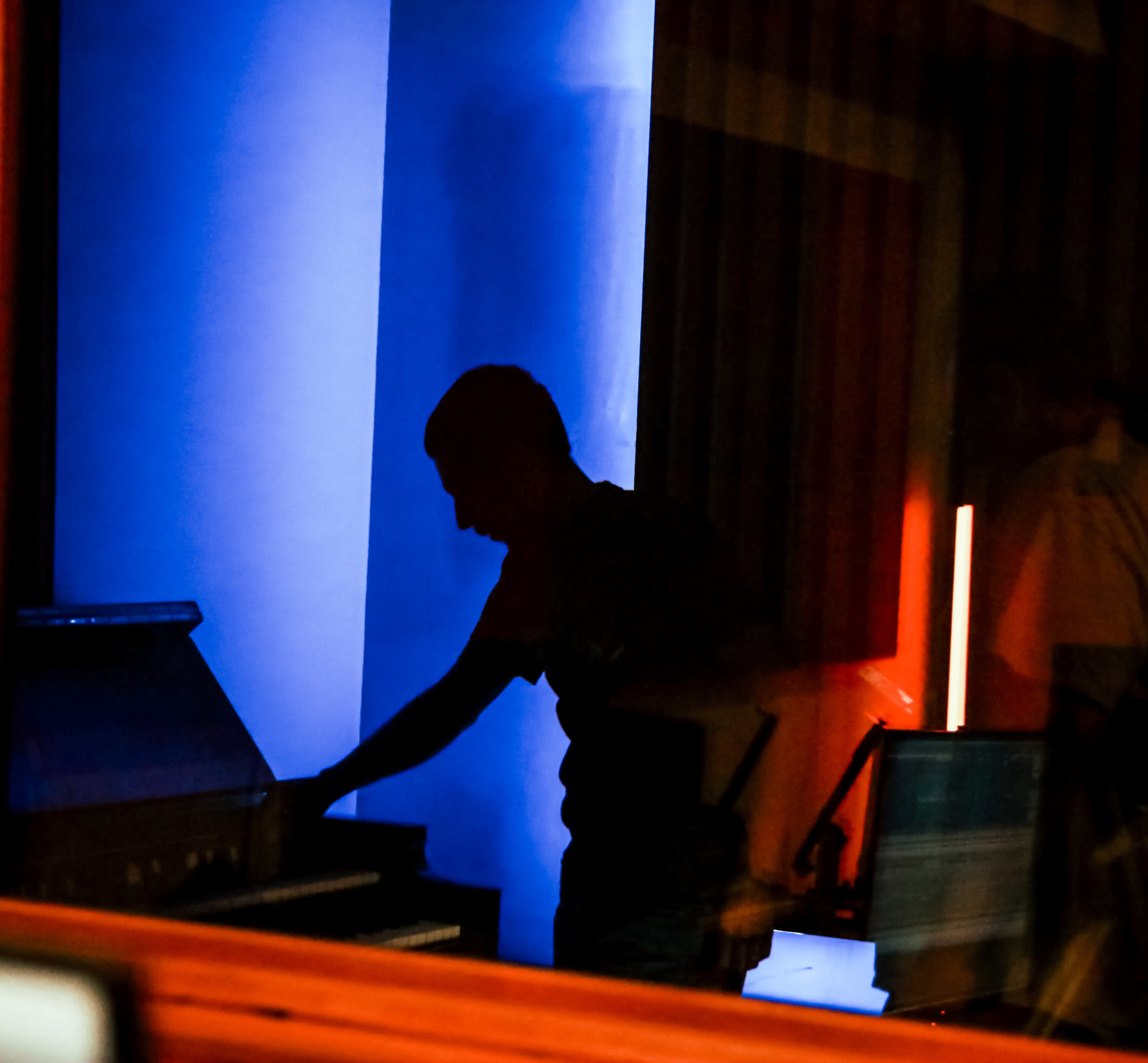 A silhouette of a person is seen reaching towards a piano in a dimly lit room with blue and orange lighting.