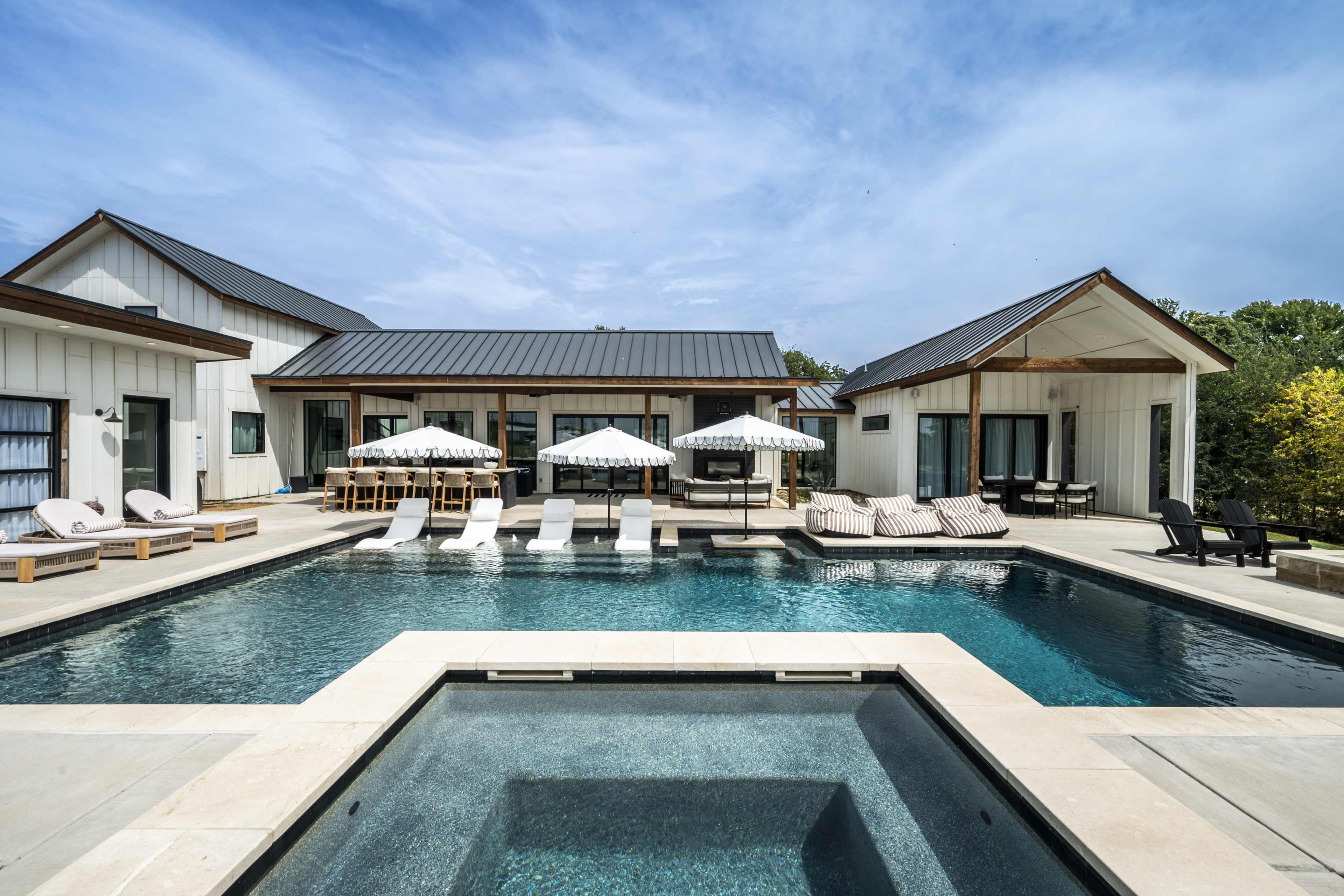 The image shows a modern pool area featuring a rectangular swimming pool, lounge chairs, and shaded umbrellas, with a stylish house in the background.
