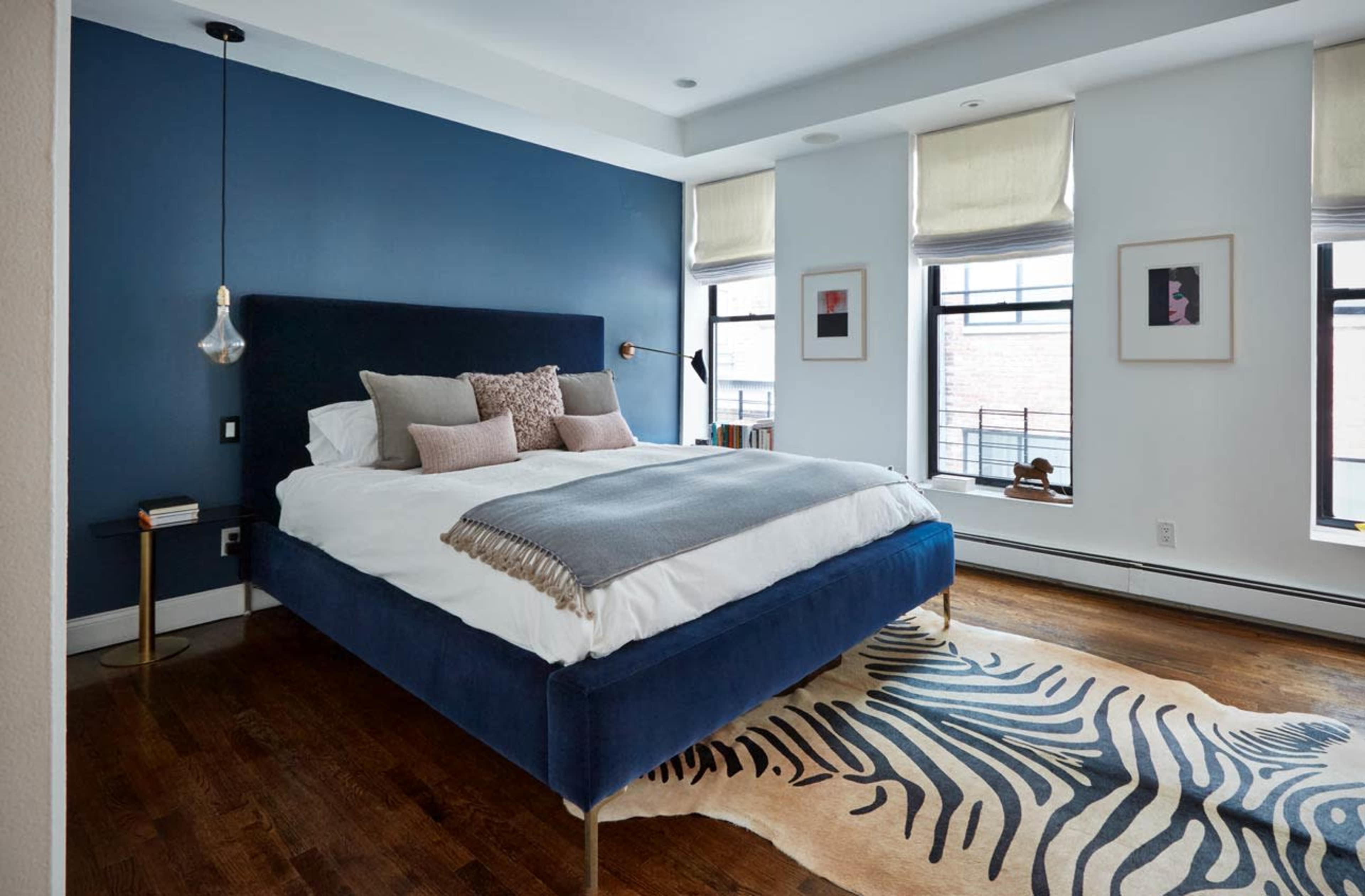 A modern bedroom features a dark blue accent wall, a large bed with decorative pillows, and a striped area rug on hardwood flooring.