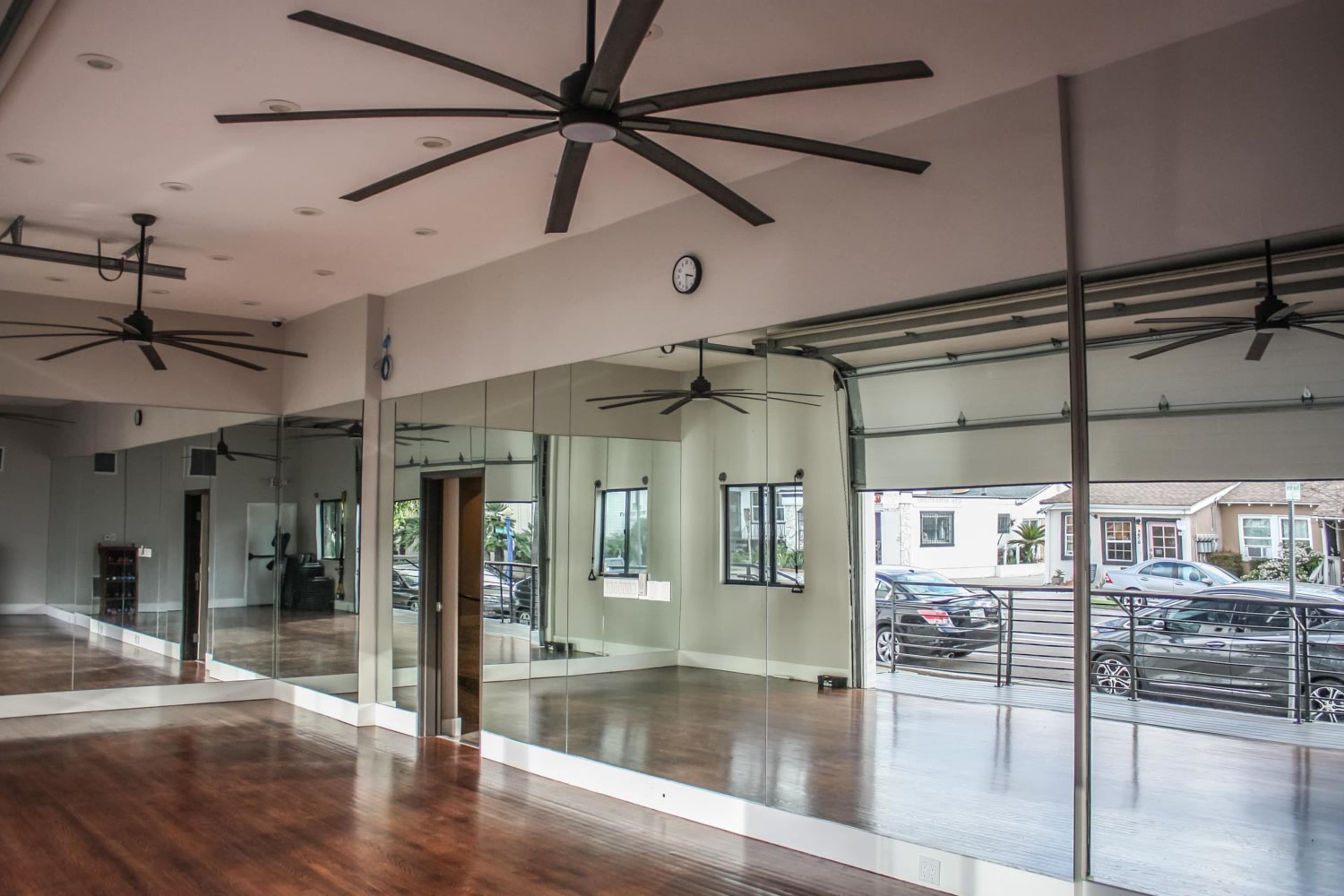 A spacious studio with wooden floors, large mirrors, and ceiling fans, featuring a large roll-up door leading to an outdoor area.