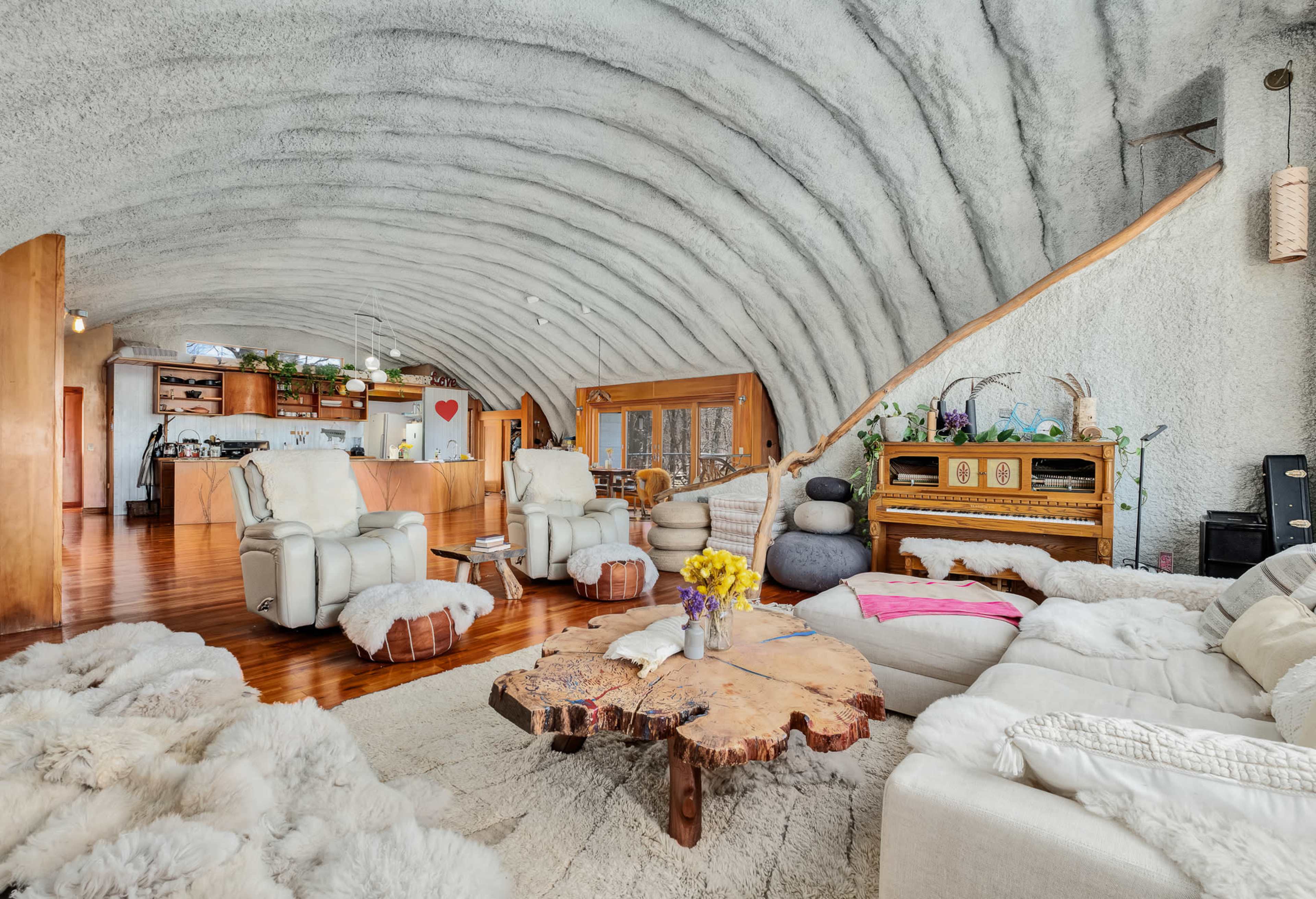 The image shows a unique living room with curved walls, two white armchairs, a wooden coffee table, and a piano in the corner, all featuring natural decor elements.