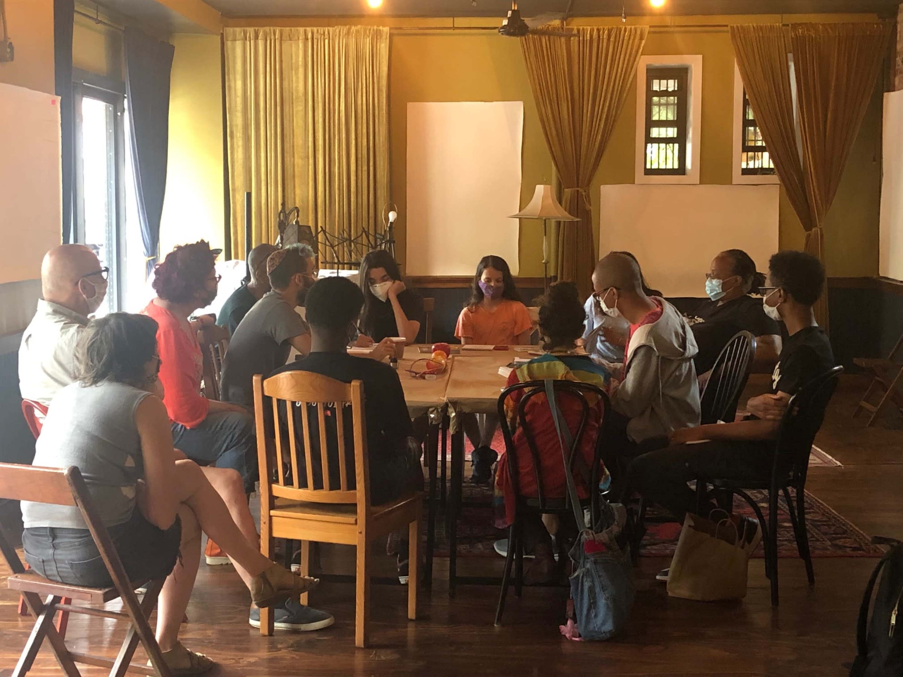 A group of people wearing masks is seated around a table in a room with yellow walls and curtains.