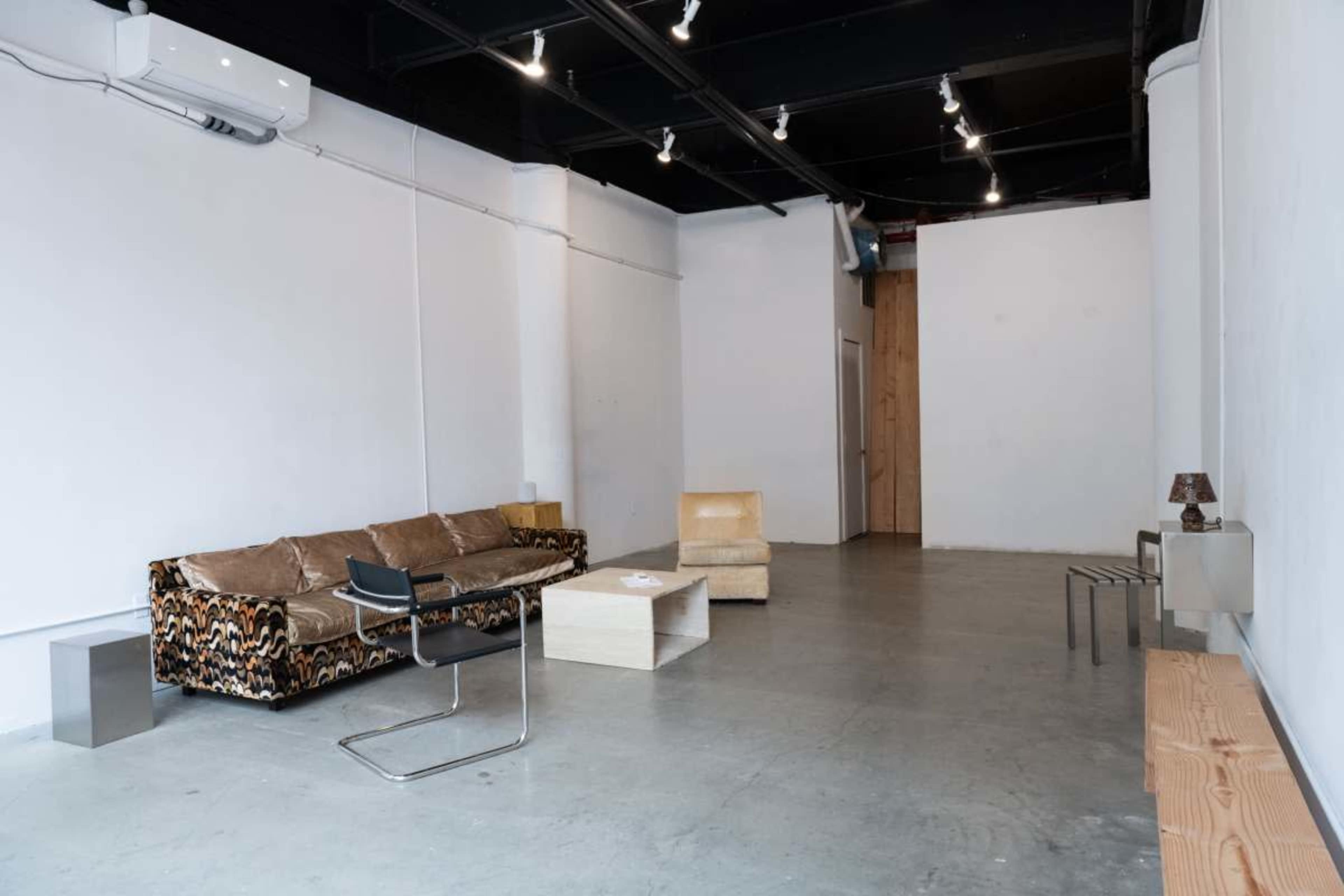 The image shows an empty interior space with a patterned sofa, a simple armchair, a small table, a chair, and a wooden console against a white wall.