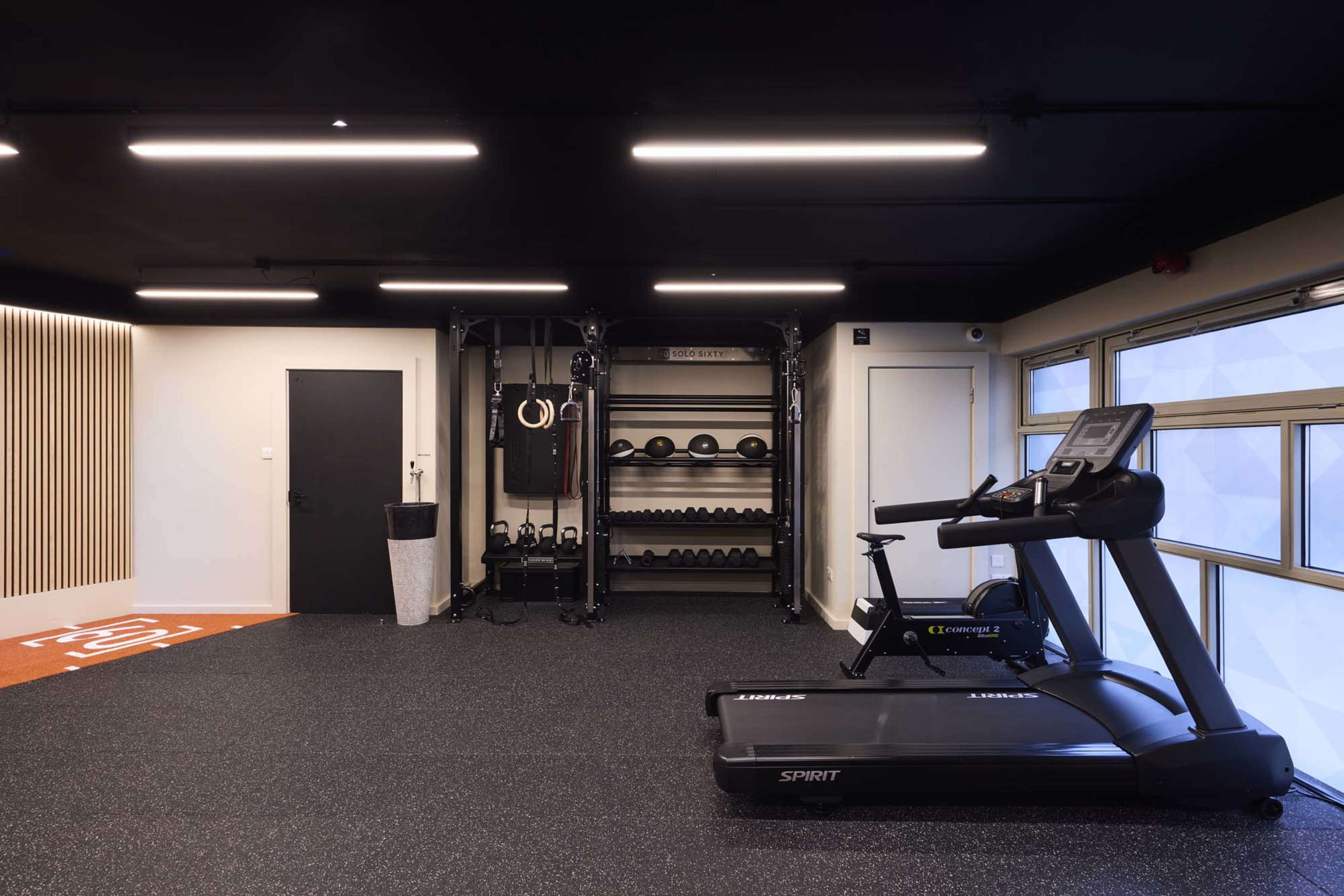The image shows a modern gym interior featuring a treadmill, a set of free weights on shelves, and exercise equipment against a dark backdrop.