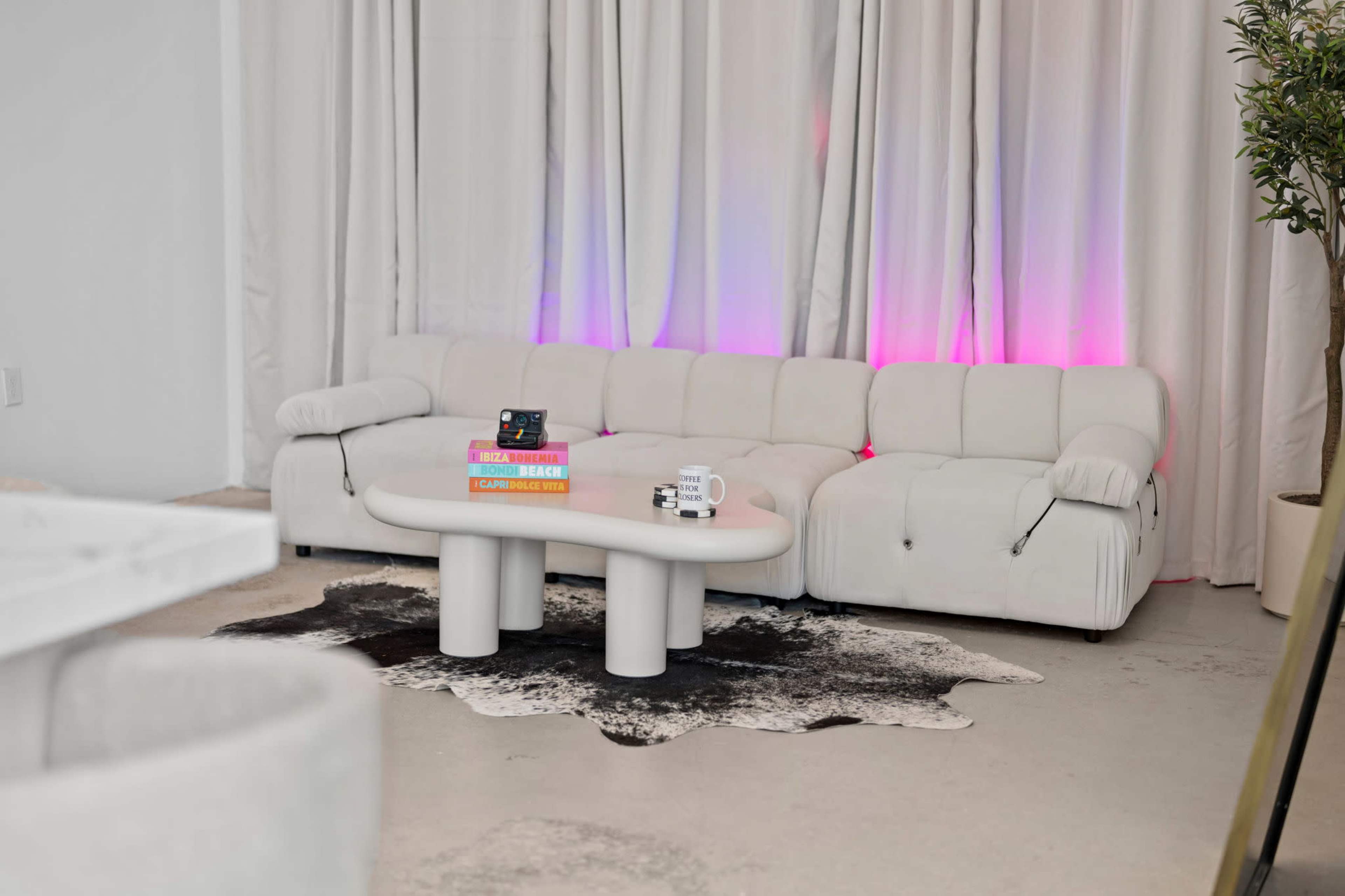 A white sectional sofa and a round coffee table are arranged in a minimalist room with curtains and soft lighting that casts a pink hue.