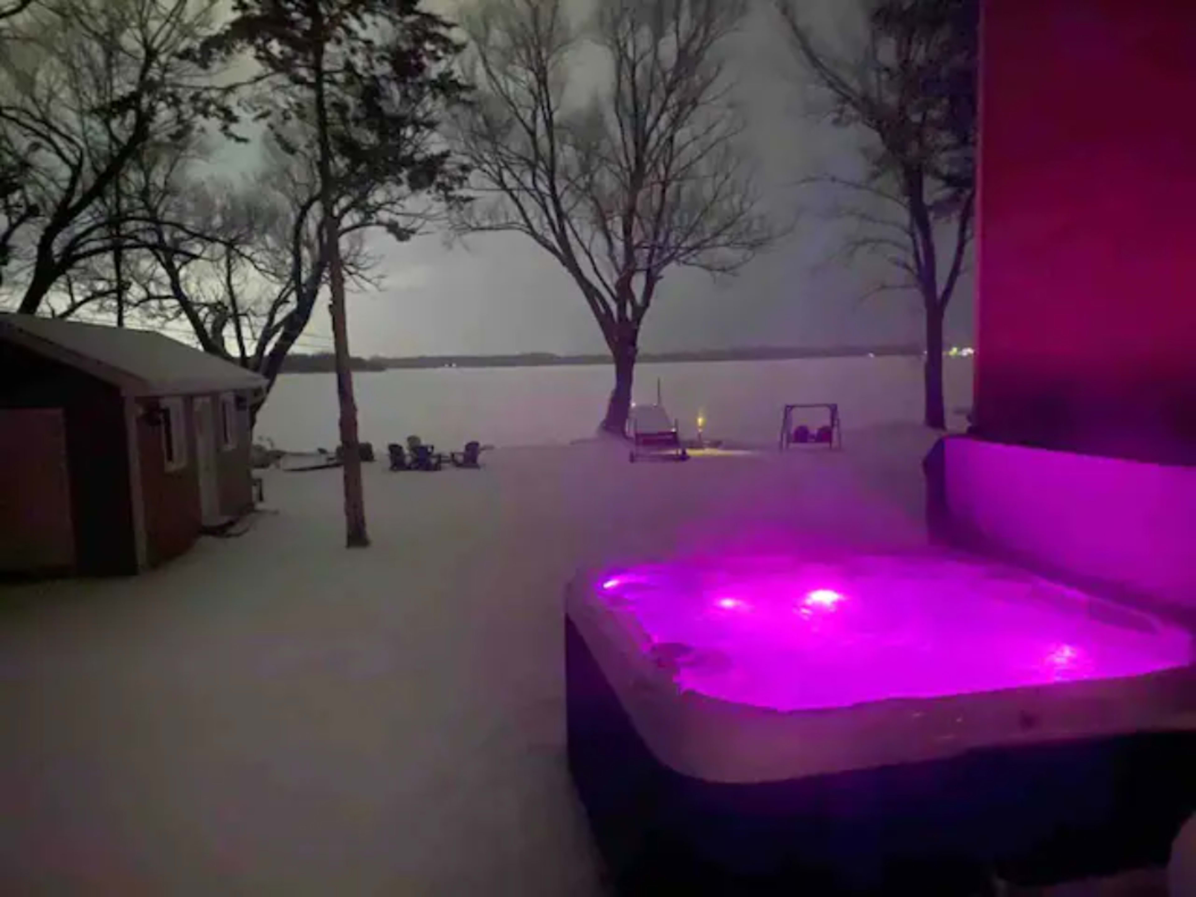 A hot tub with purple lights sits in the snowy yard near a lake, framed by leafless trees.