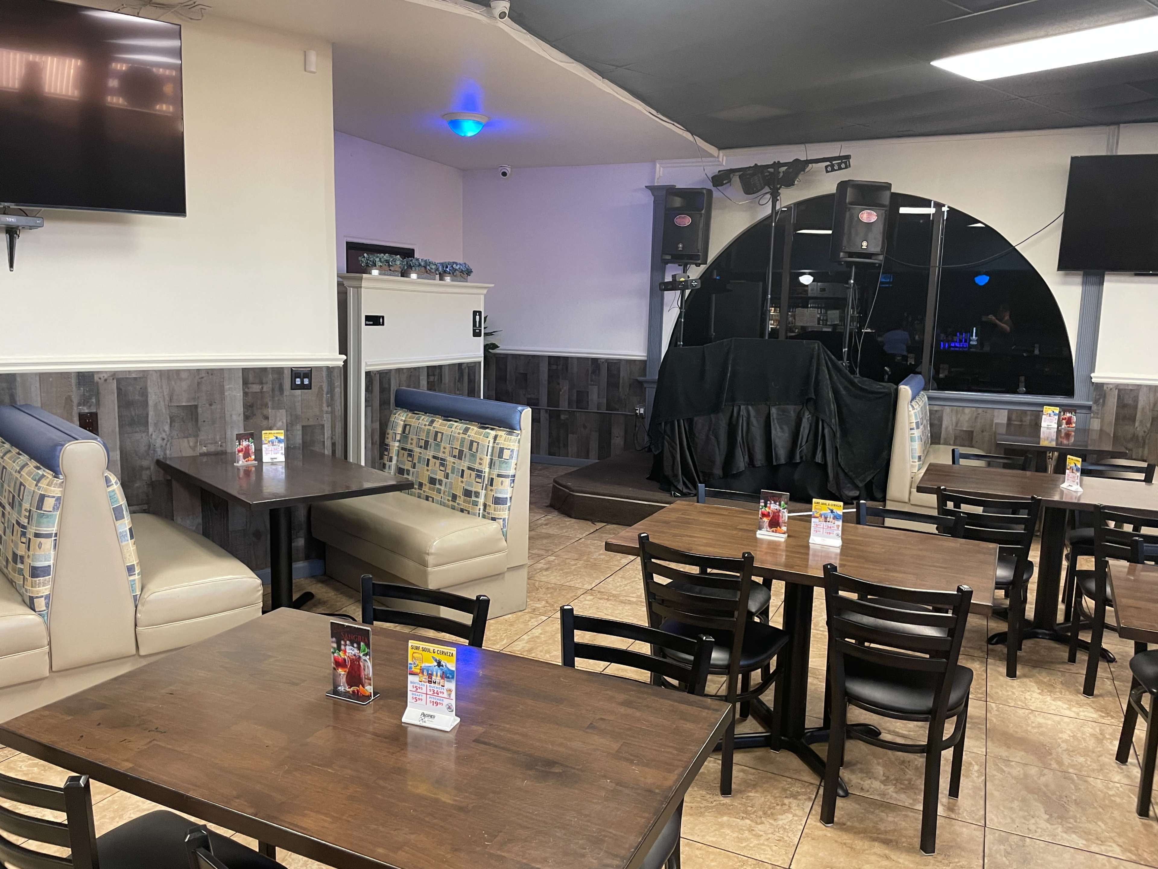 An empty restaurant interior with booth seating and tables arranged in a casual dining layout, featuring a small stage and audio equipment in the background.