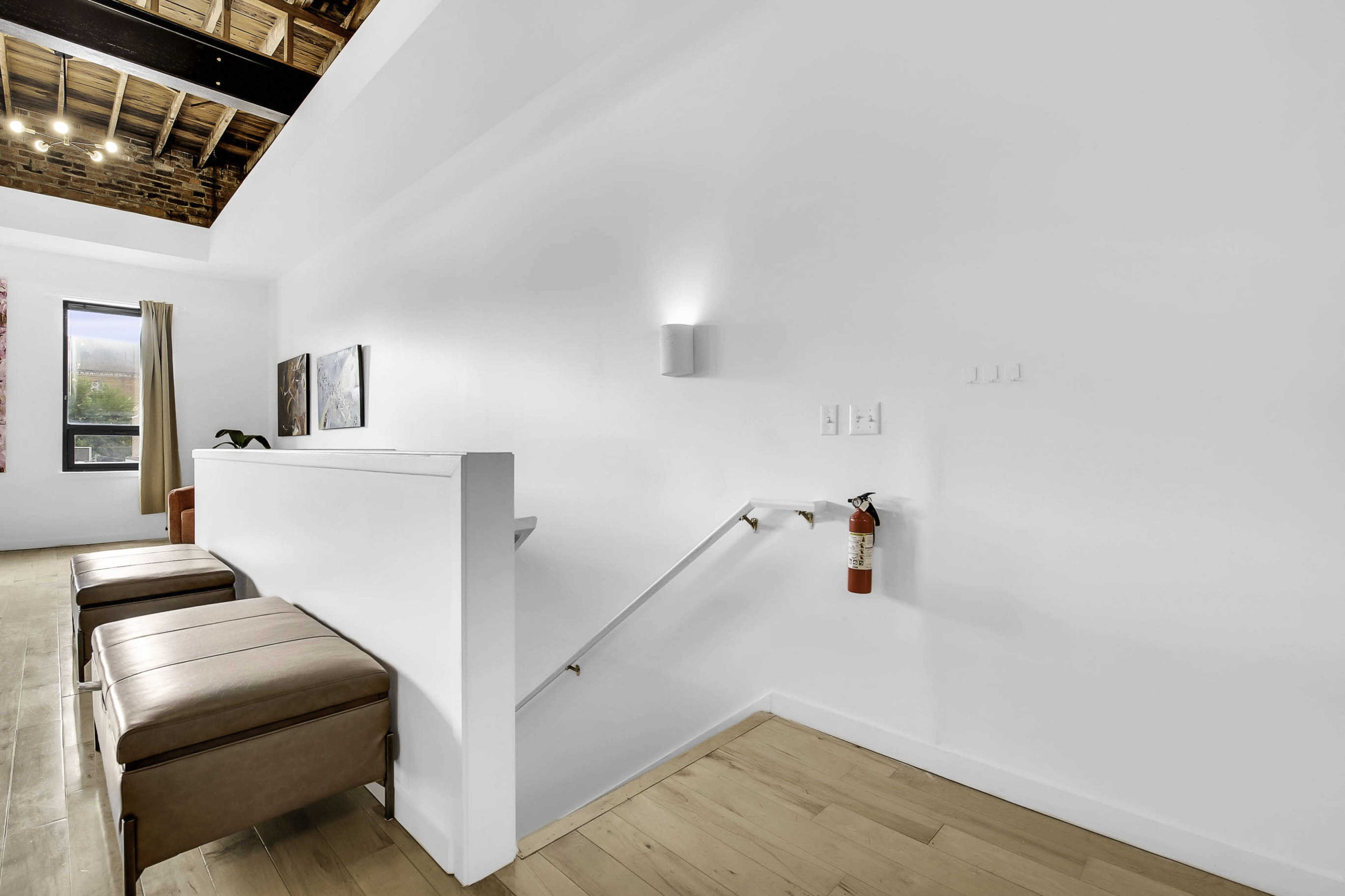 A minimalist interior with a white wall, wooden flooring, and a simple staircase accompanied by two leather stools.