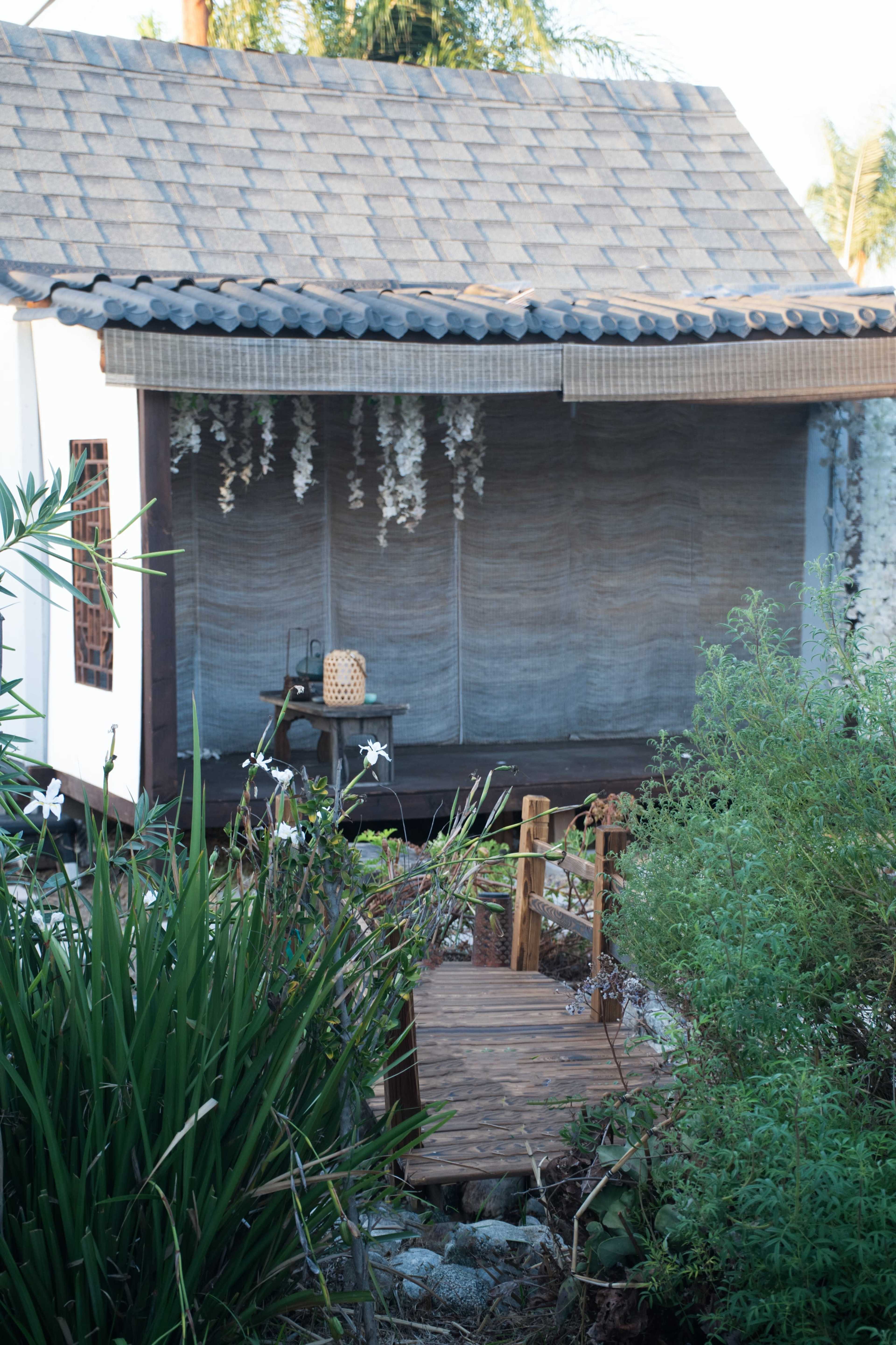 A small wooden bridge leads to a house with a shingled roof, surrounded by greenery and hanging decor.