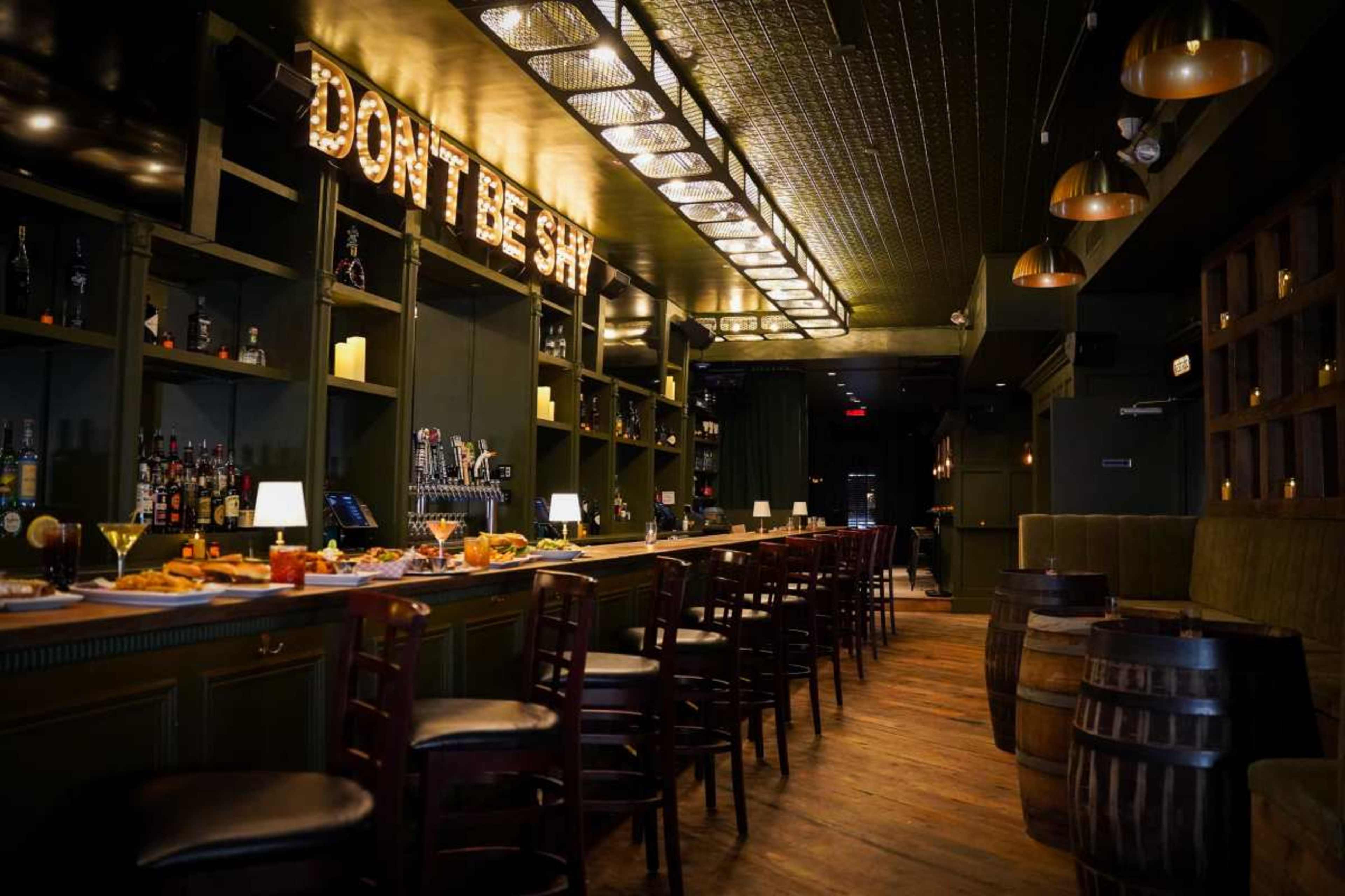 A dimly lit bar features a long wooden counter lined with high stools, shelves stocked with liquor, and a variety of appetizers displayed on the surface.