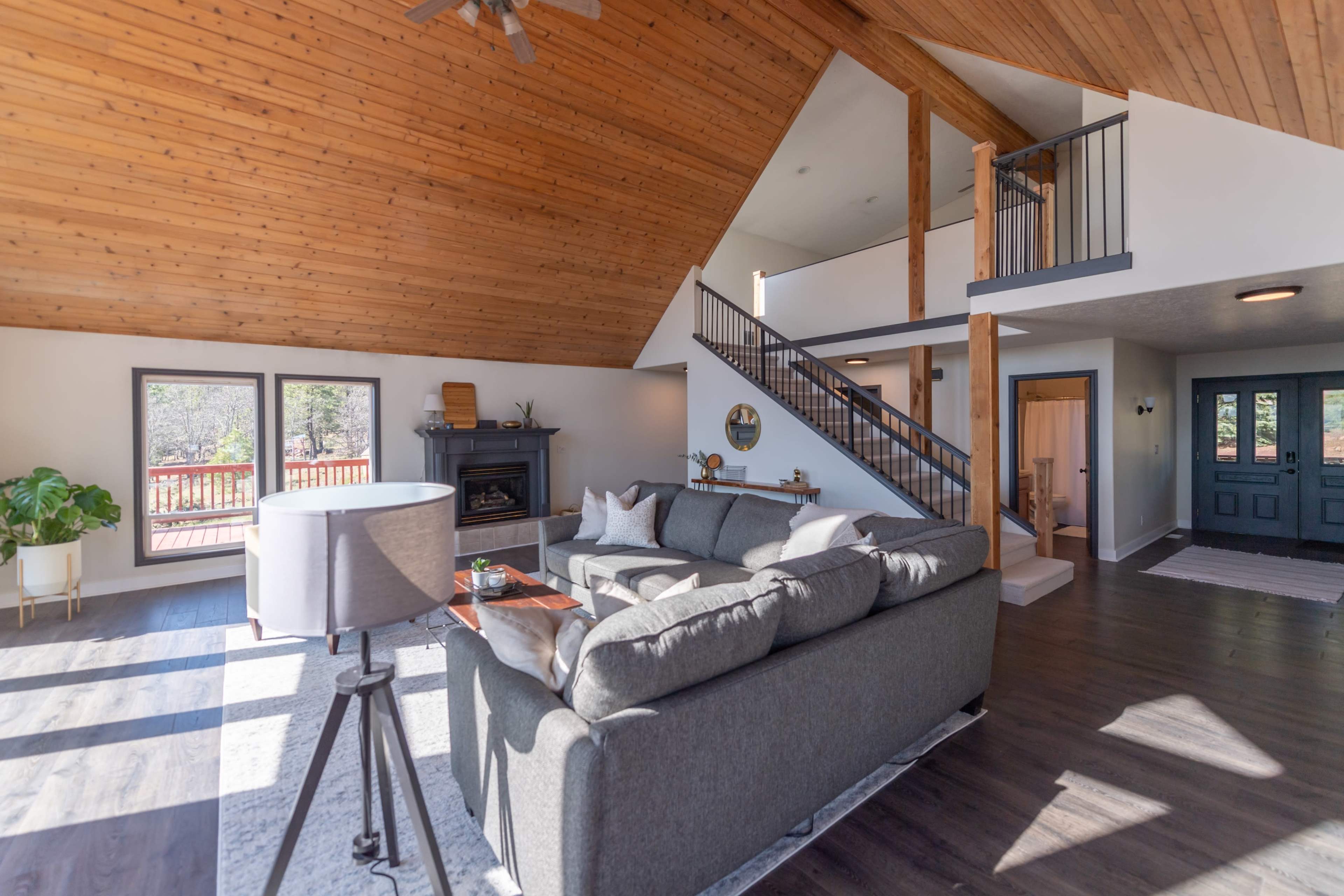 The living room features a spacious layout with a high wooden ceiling, large windows, a gray sectional sofa, and a staircase leading to an upper level.