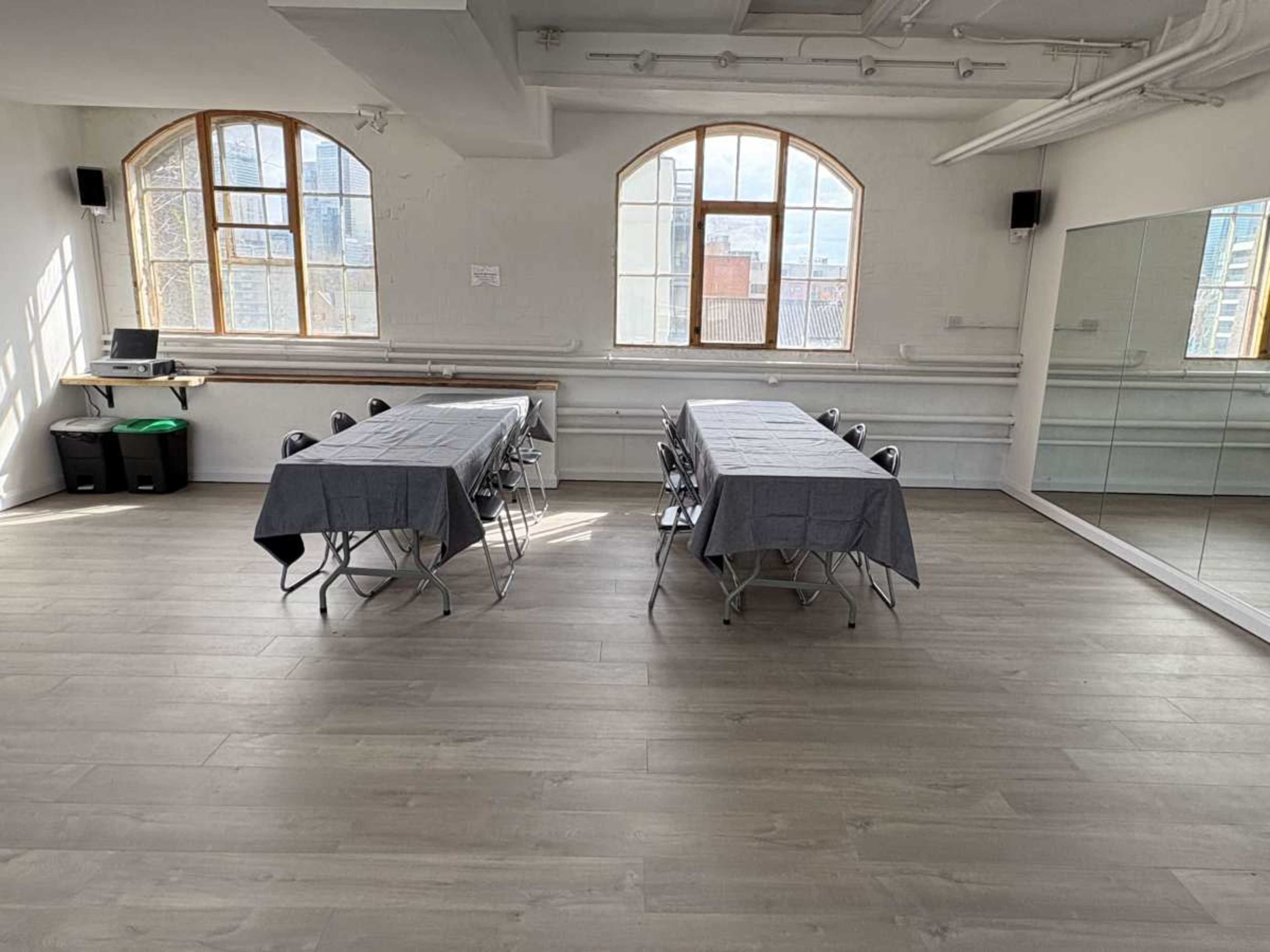 The image shows a bright, spacious room with two tables covered in gray tablecloths, placed parallel to each other, and large windows allowing natural light to enter.