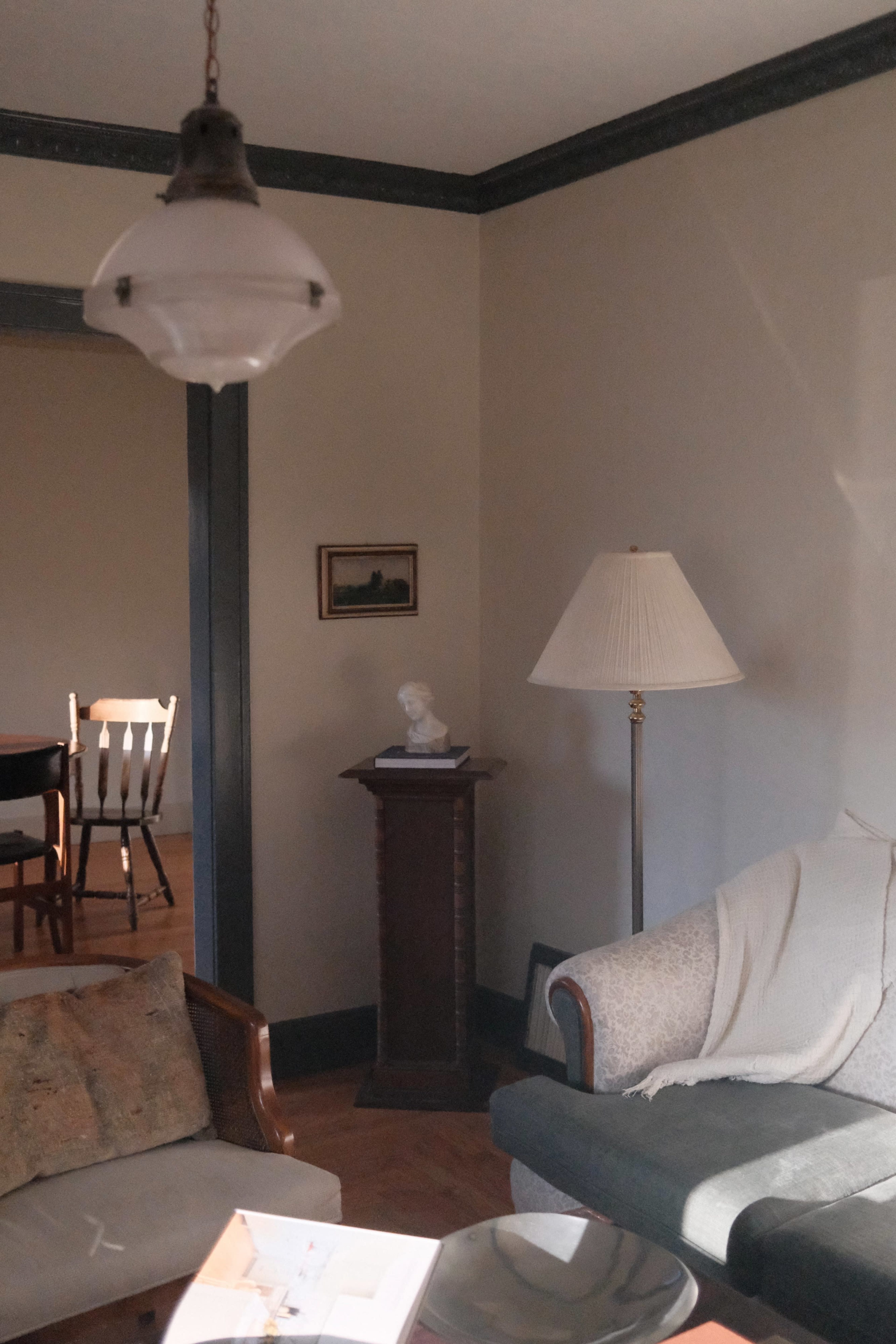 A softly lit living room with a sofa, a side table, a floor lamp, and a small bust on a pedestal, with a dining area visible in the background.