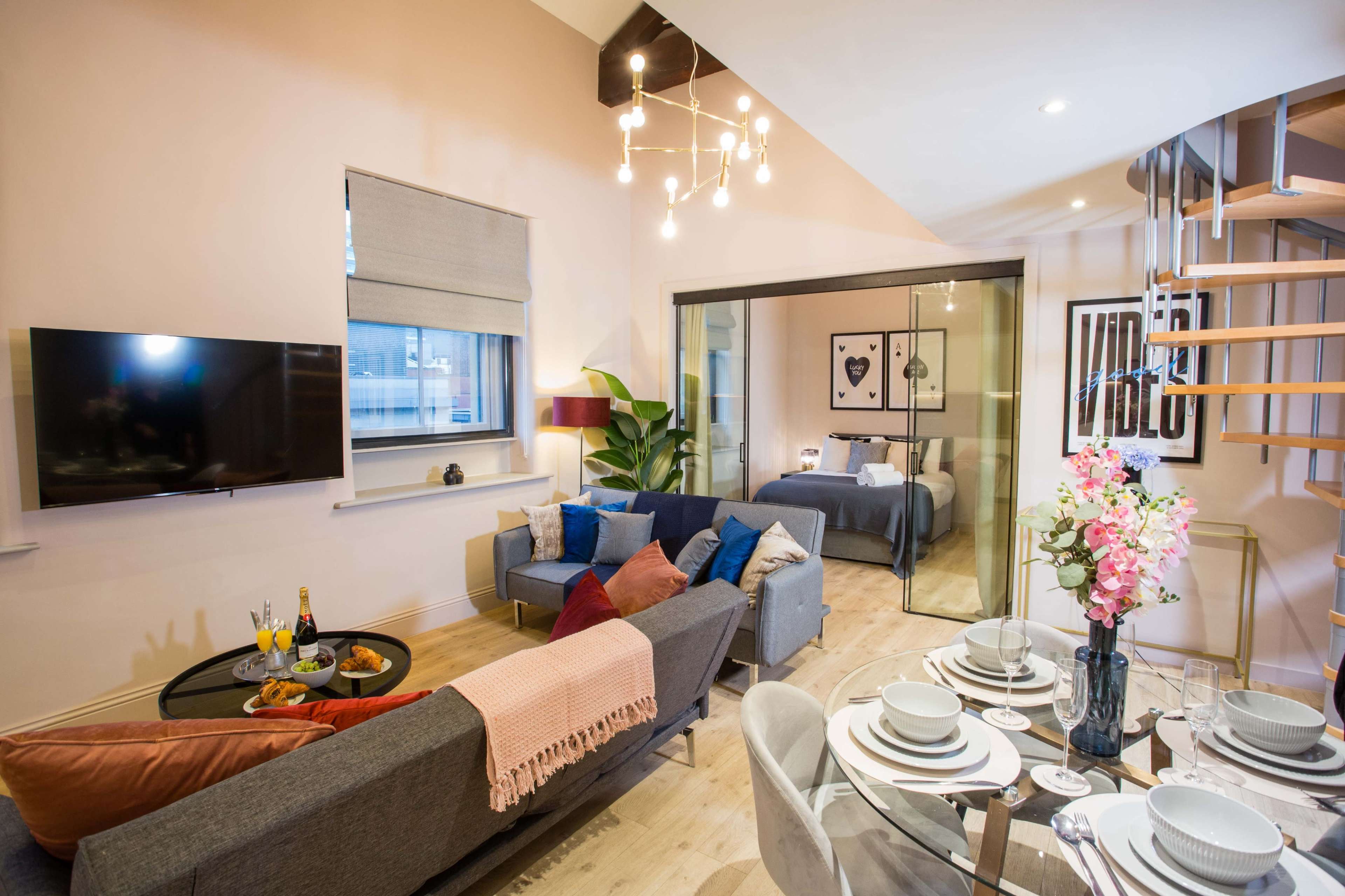 The image shows a modern living space featuring a gray sofa, a glass dining table set for a meal, and a staircase leading to a bedroom area.
