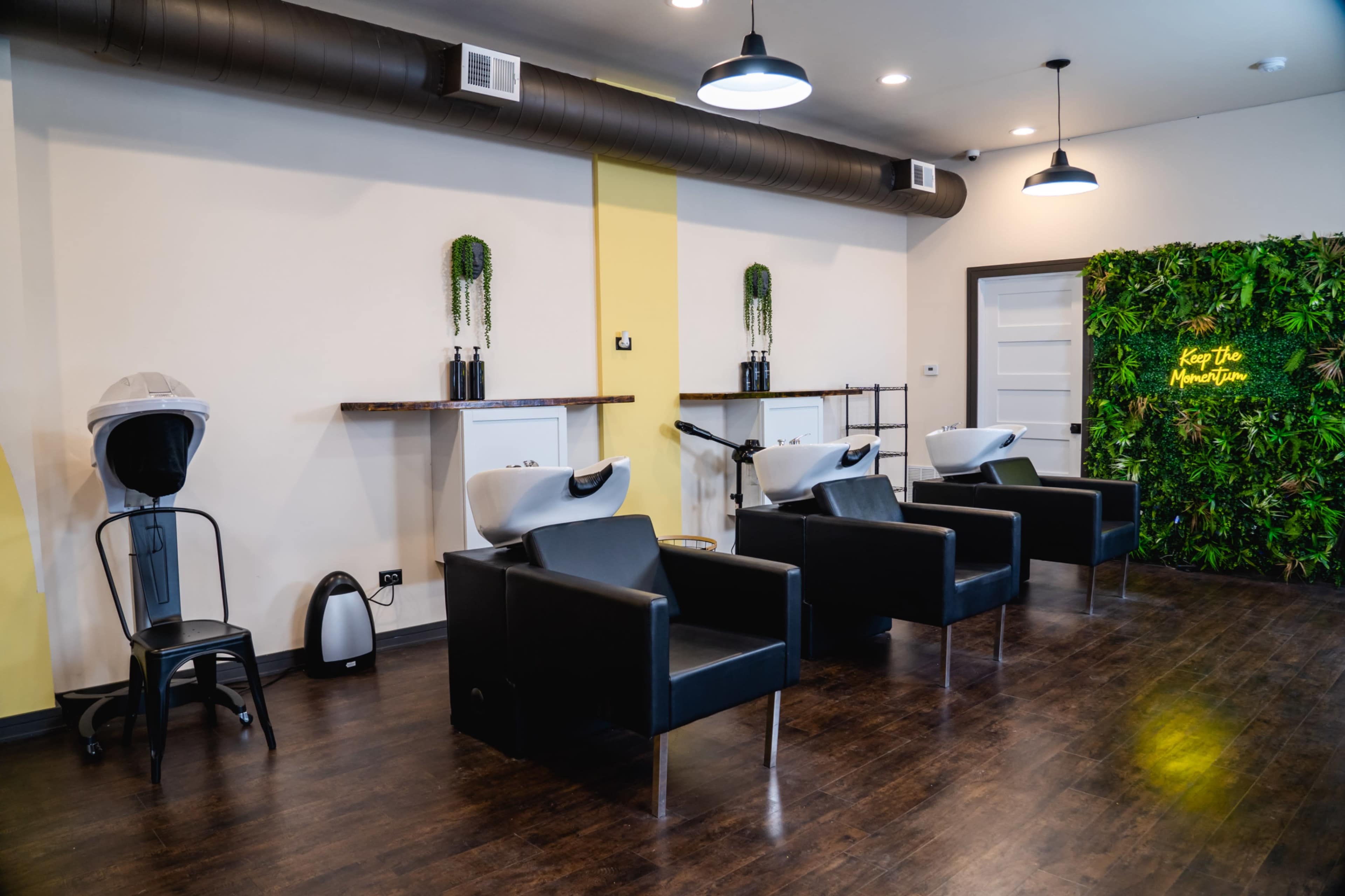 A modern salon interior featuring four black salon chairs and two shampoo stations against a wall adorned with greenery.