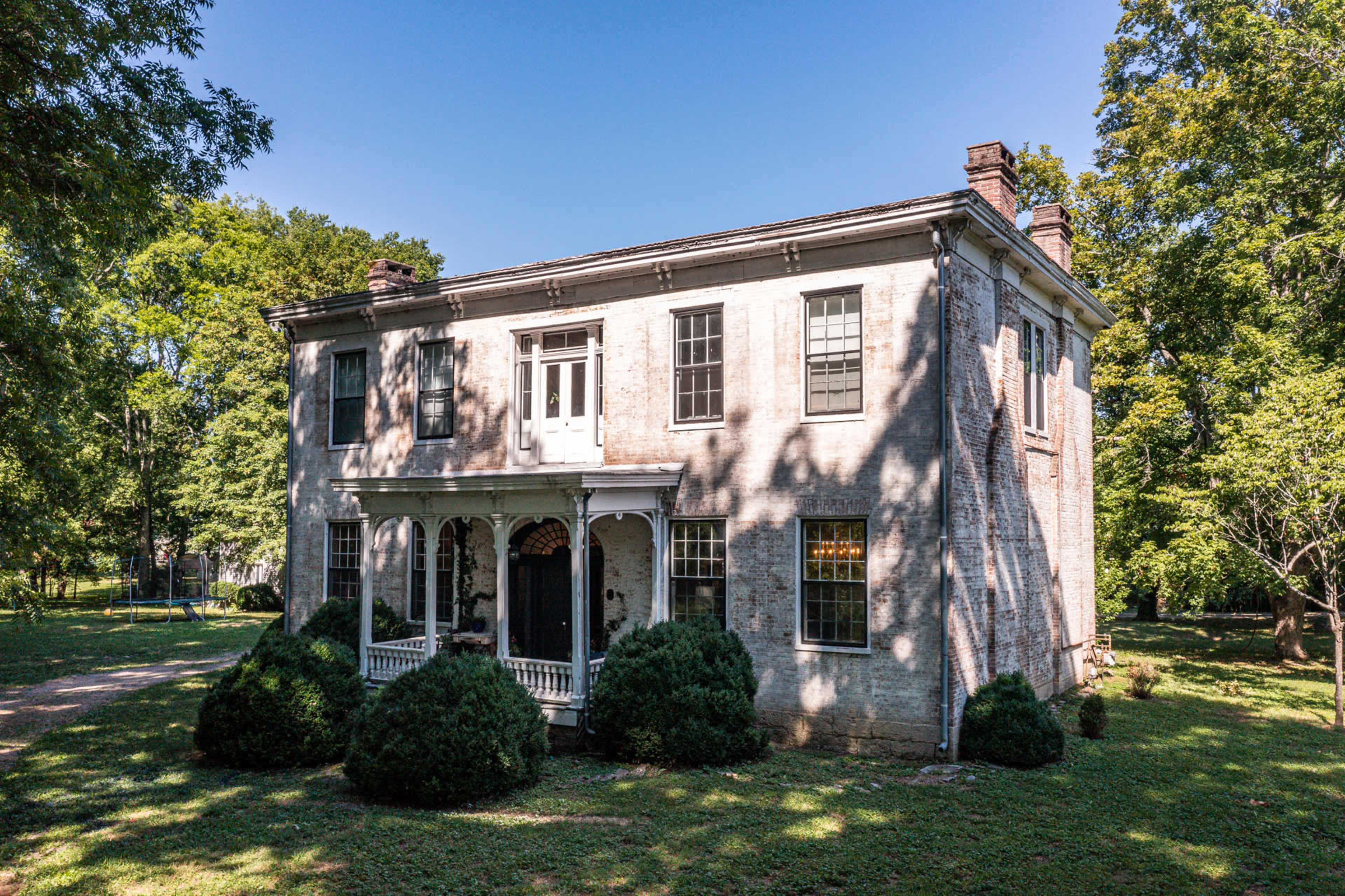 A two-story brick house with a front porch surrounded by manicured shrubs and trees.