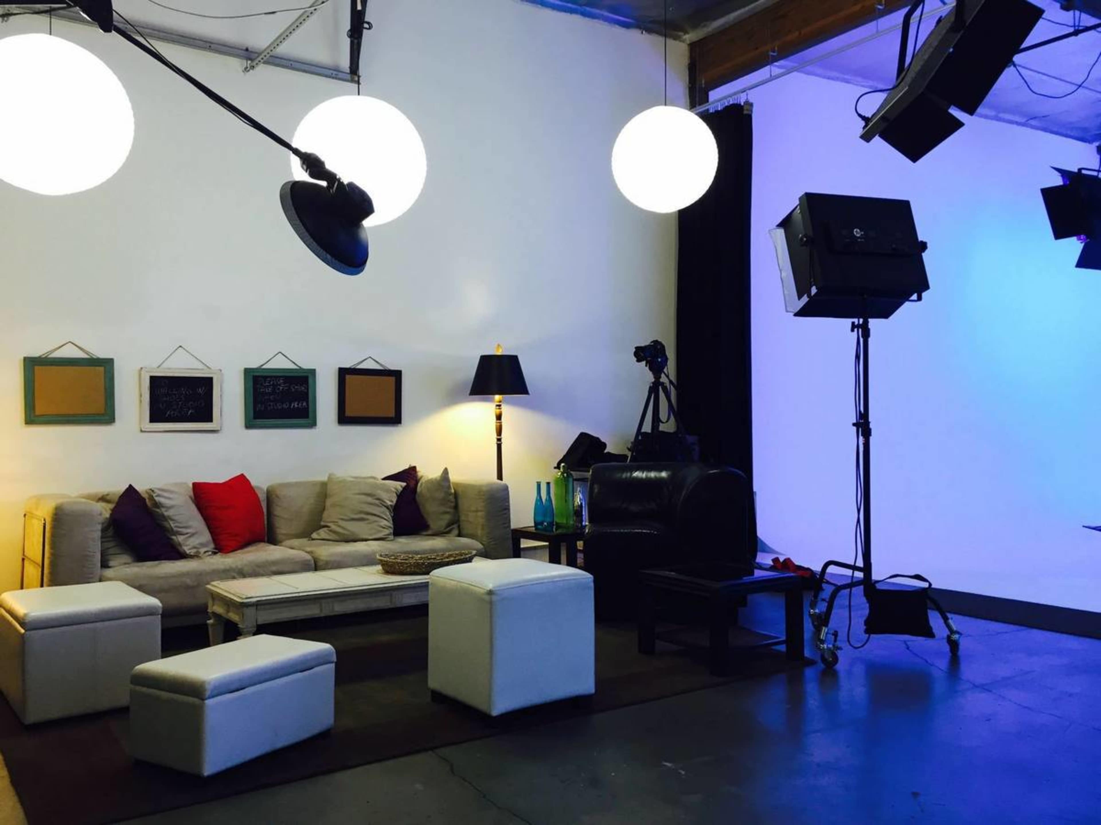 A modern studio setup with a beige couch, white ottomans, a floor lamp, and photography equipment against a backdrop of a blue-lit wall.