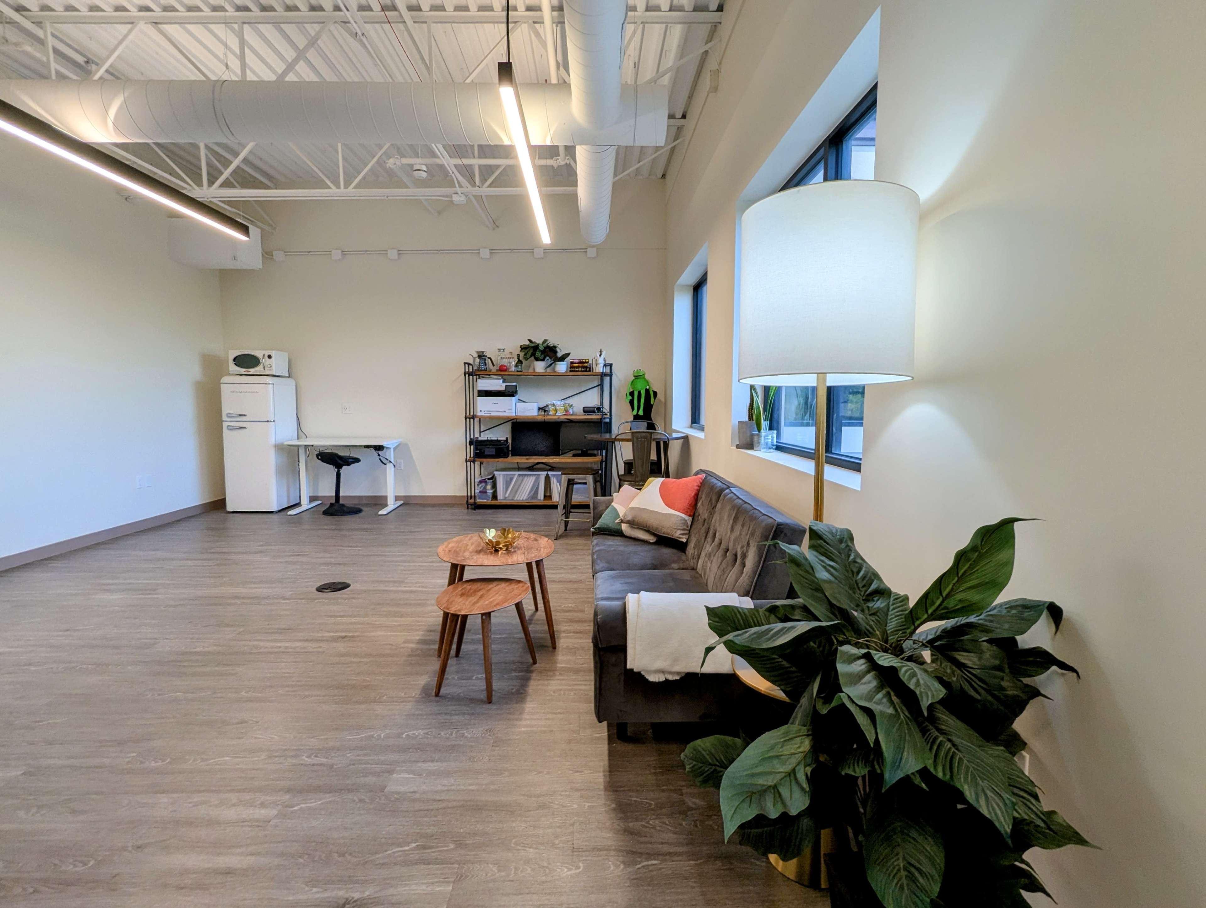 A modern interior space features a grey sofa, a small wooden coffee table with a decorative item, a floor lamp, and a potted plant, with a kitchenette and shelves visible in the background.