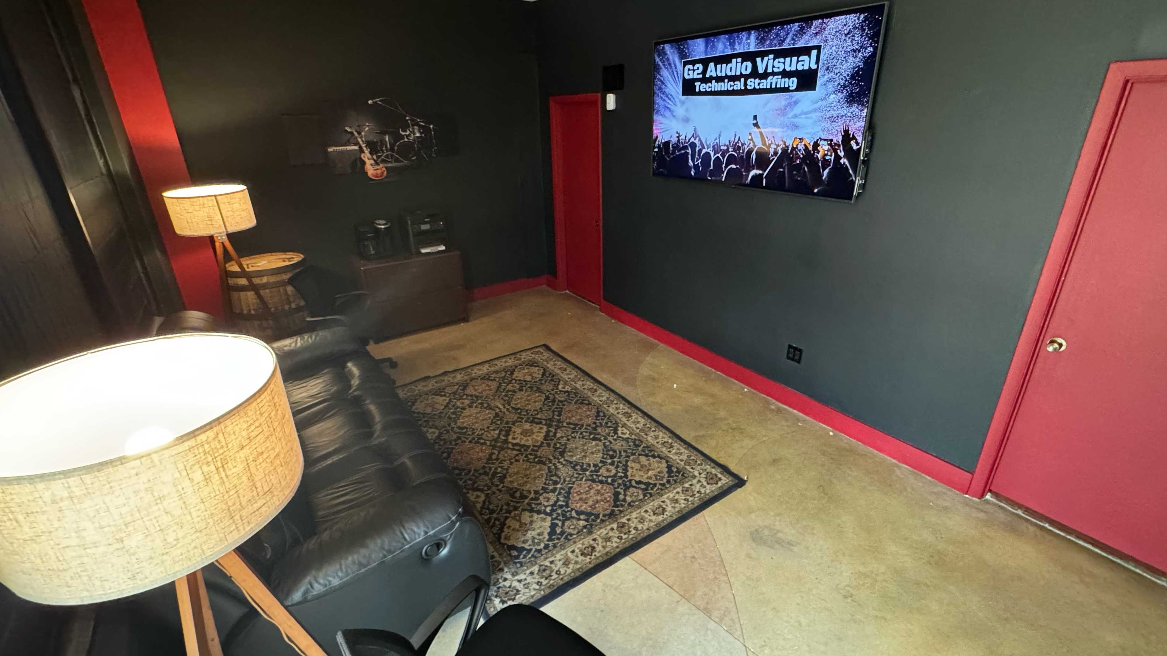 The image shows a small, dimly lit room with black walls, a large television displaying "G2 Audio Visual Technical Staffing," a patterned rug, a lamp, and leather seating.
