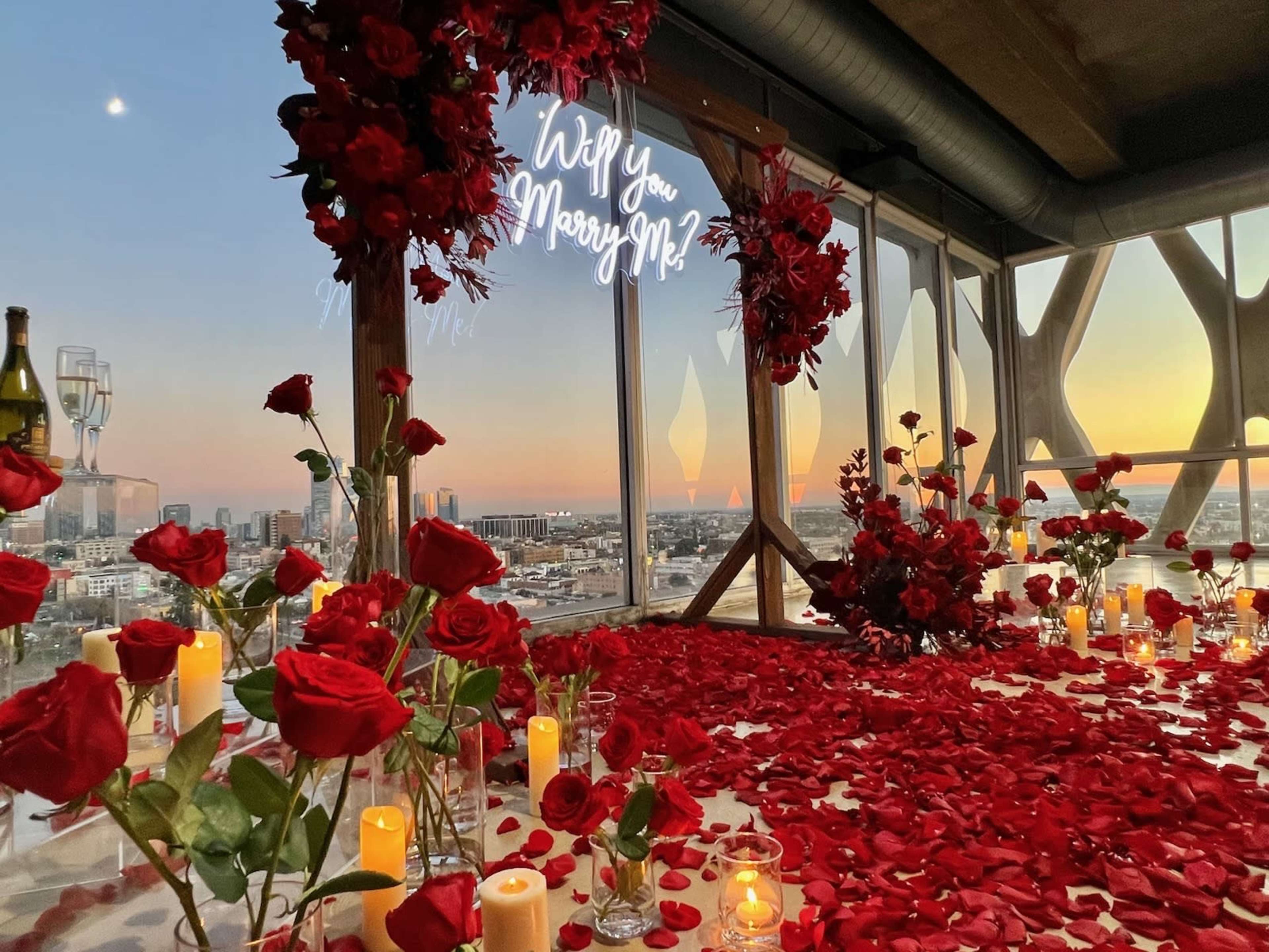 A romantic proposal setup features a floor covered in rose petals, surrounded by candles, with a glowing sign that reads "Will you Marry Me?" and a city skyline visible through large windows.