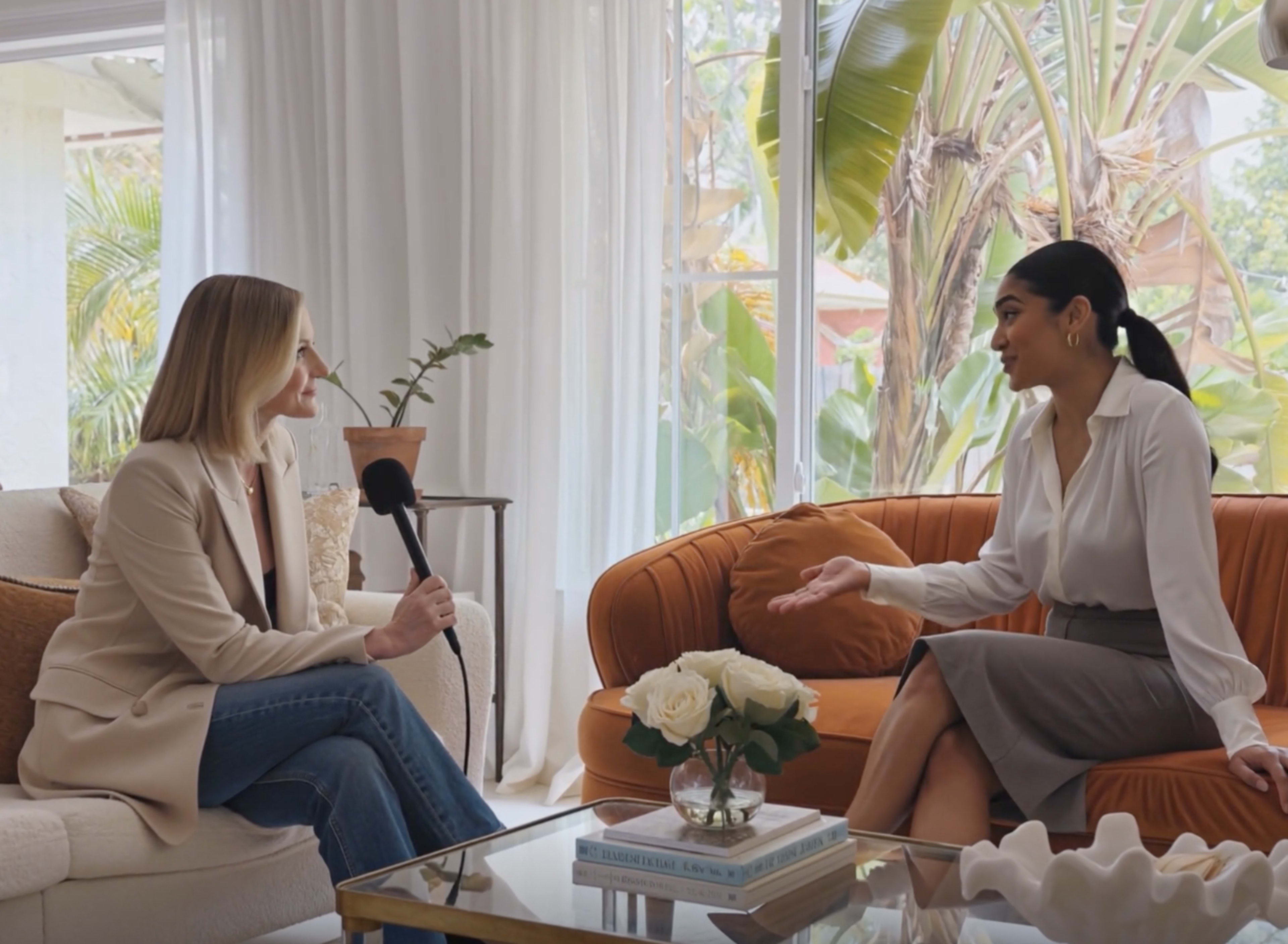 Two women are seated in a well-lit living room, engaging in conversation, with a large window and greenery visible in the background.