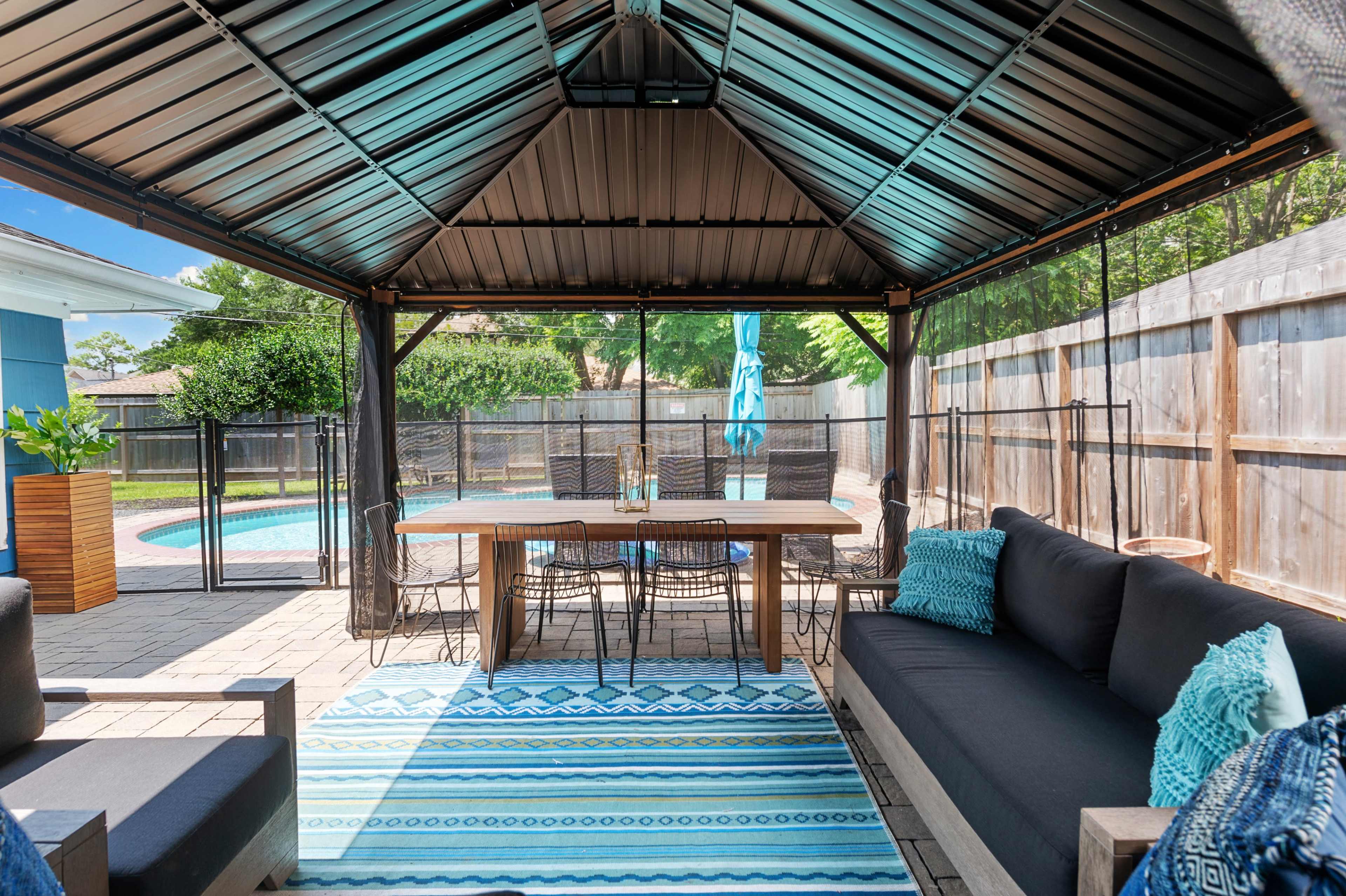 A covered outdoor seating area features a table and chairs surrounded by a pool and a fenced backyard.