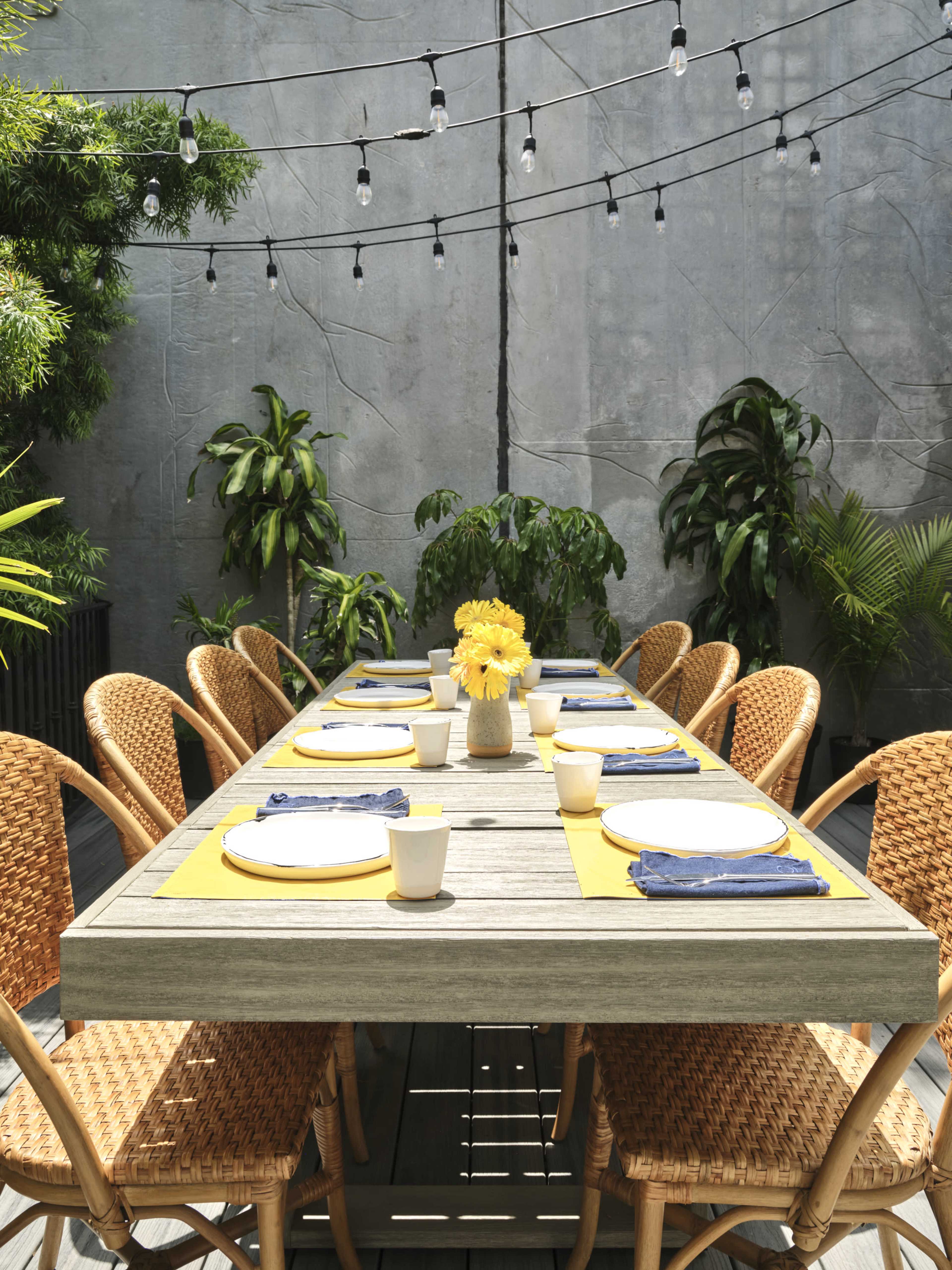 A long wooden table set for dining is surrounded by woven chairs, with a vase of yellow flowers at the center and string lights overhead in a green courtyard.