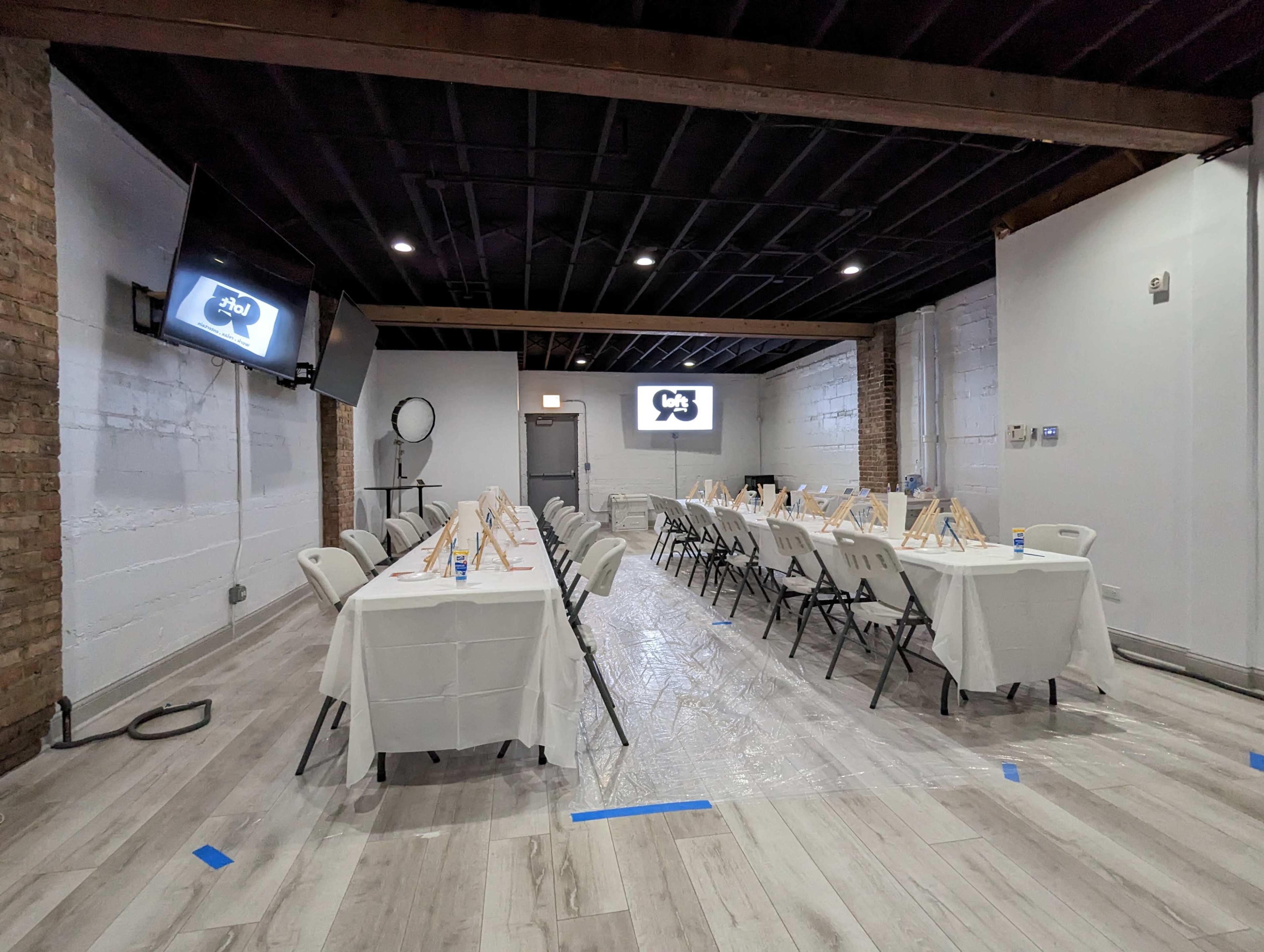 A spacious room arranged with tables covered in white tablecloths, each holding an easel, and featuring screens and a circular light setup along the walls.