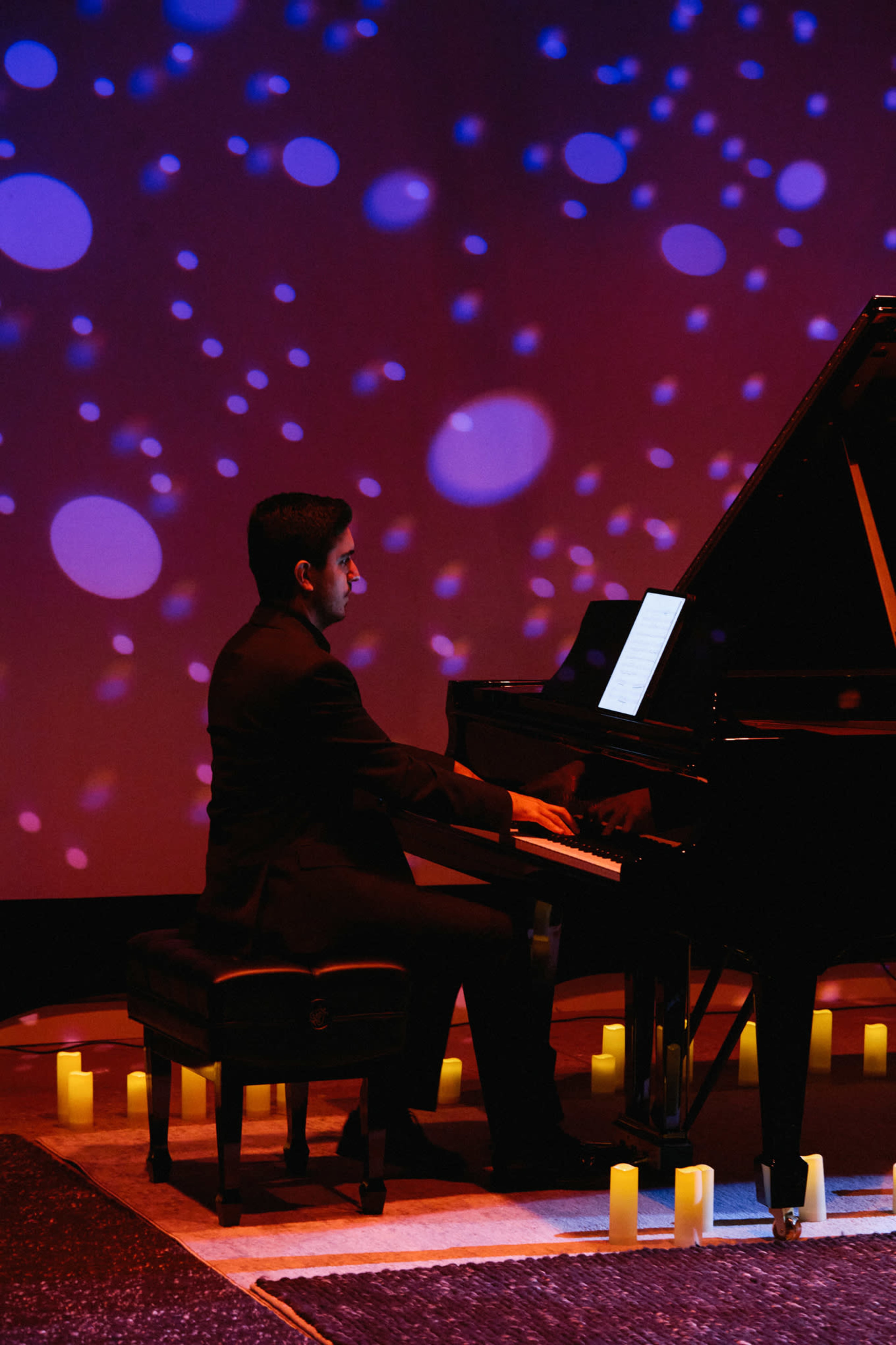 A pianist plays on a grand piano surrounded by candles, with a colorful light display in the background.