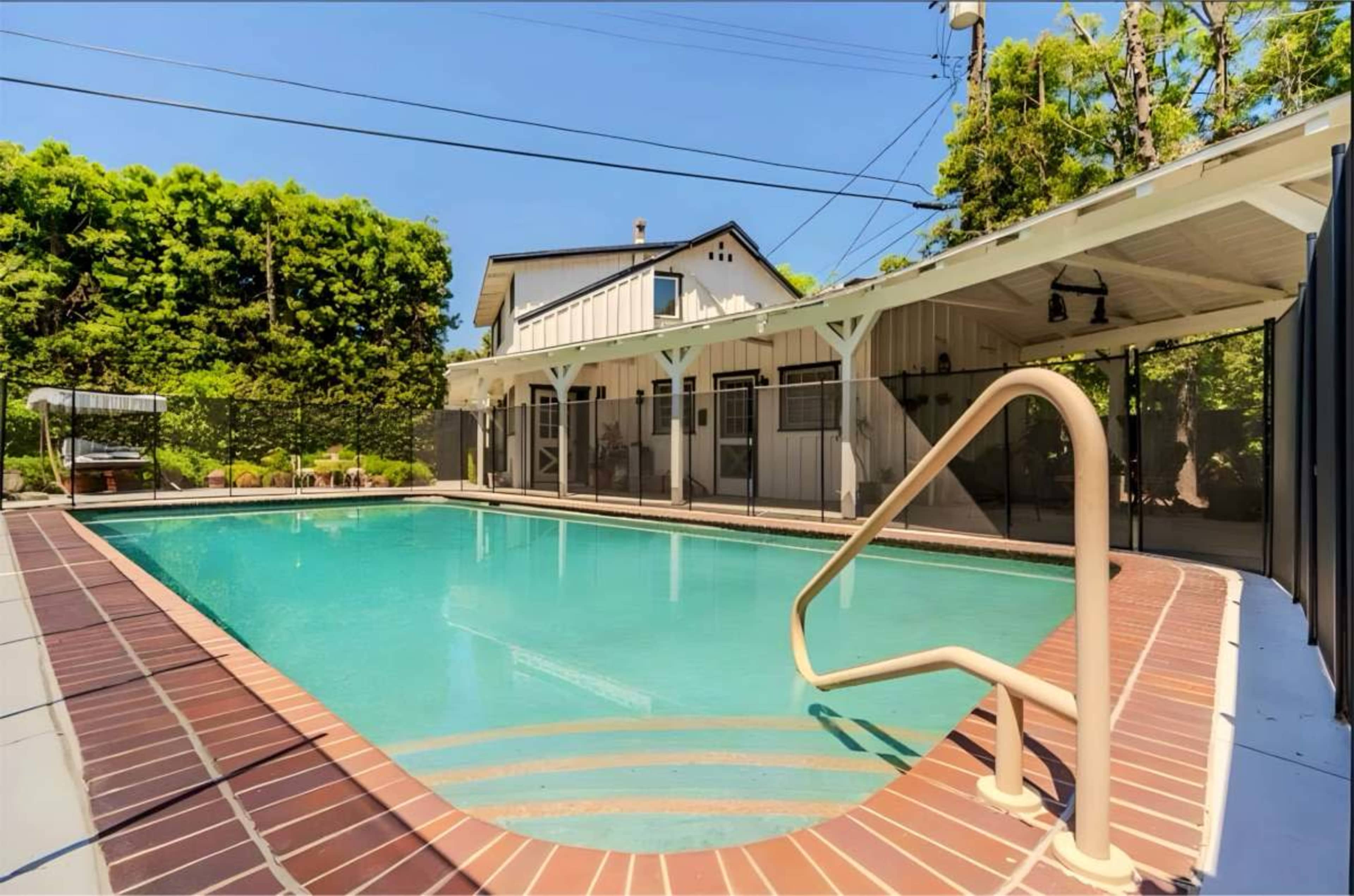 A rectangular swimming pool is surrounded by a tiled deck and is located next to a house with several adjacent patio spaces.