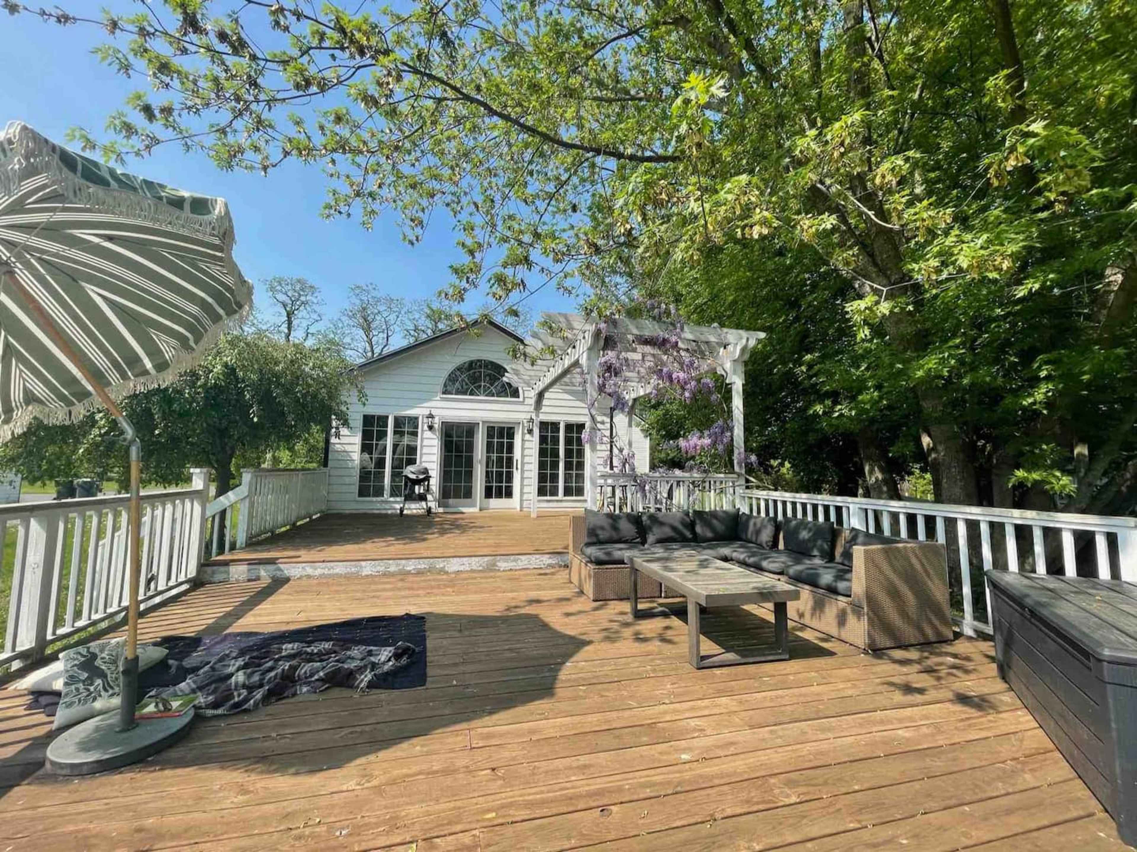 A wooden deck features outdoor seating and a large umbrella, surrounded by lush greenery and a white house with multiple windows.