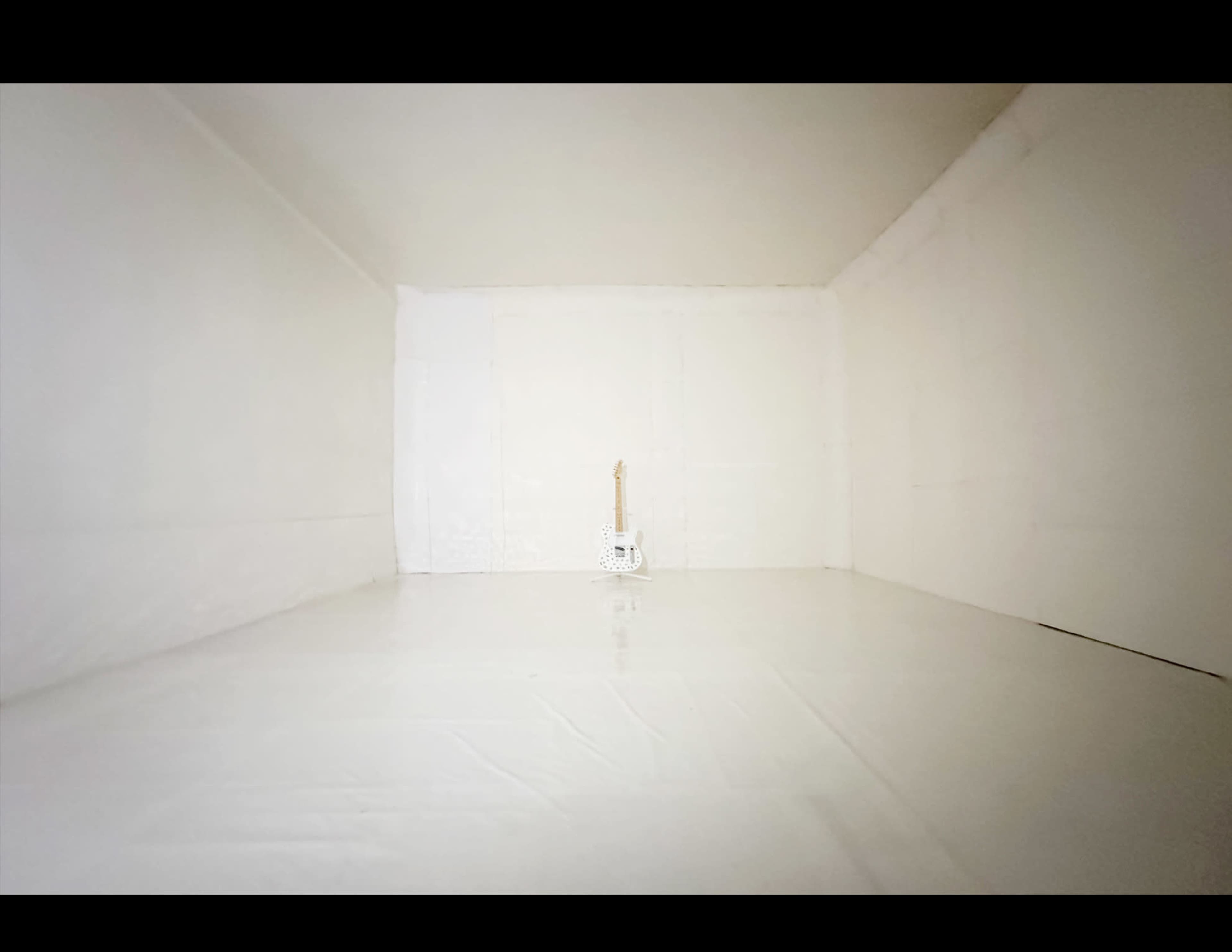 A white room with blank walls and a shiny floor contains a single electric guitar standing upright in the center.