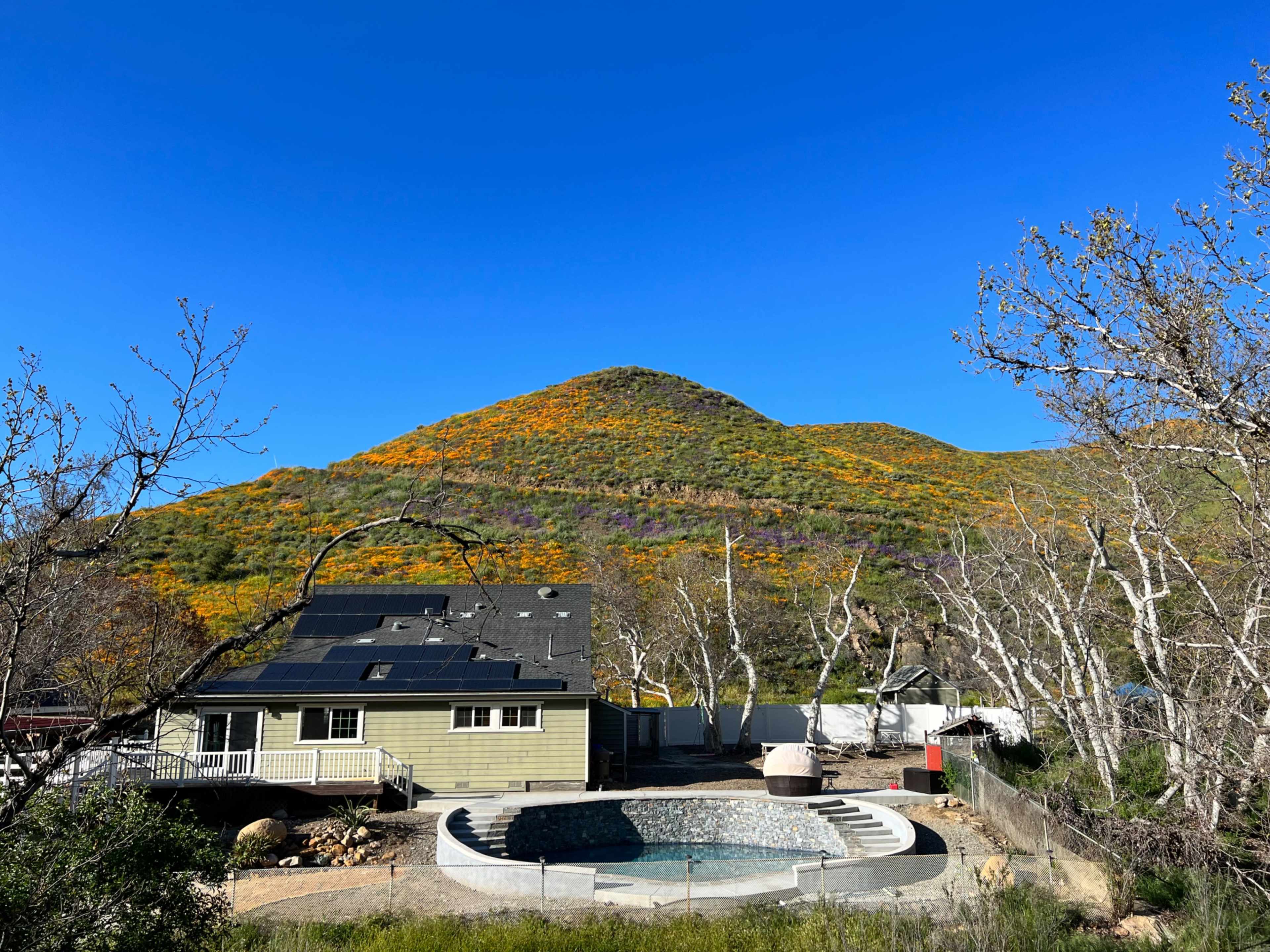 A house with a pool is situated in the foreground, while a hill covered in orange and green foliage rises in the background against a clear blue sky.