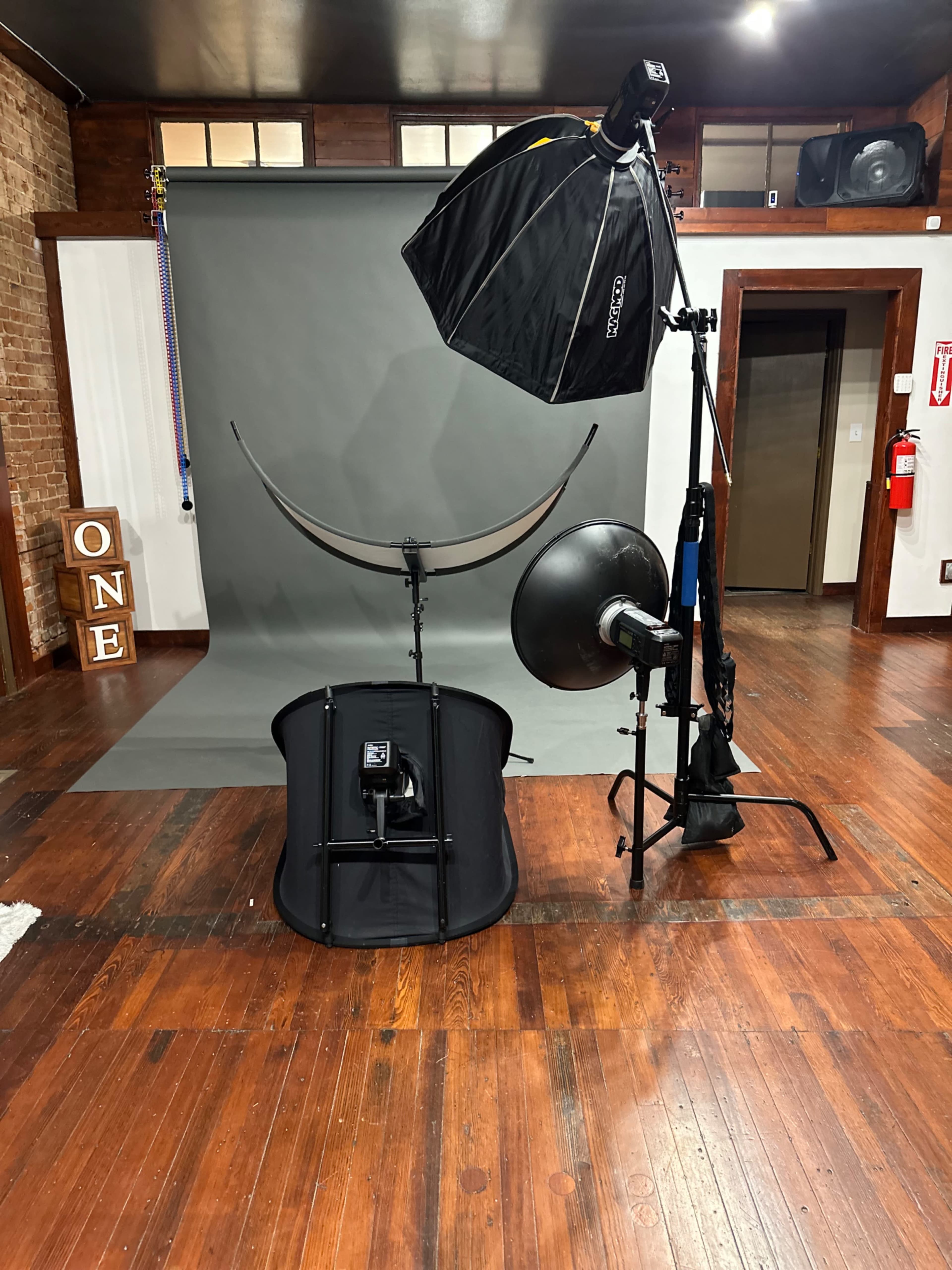The image shows a photography studio setup with a backdrop, light modifiers, and various lighting equipment arranged on wooden flooring.