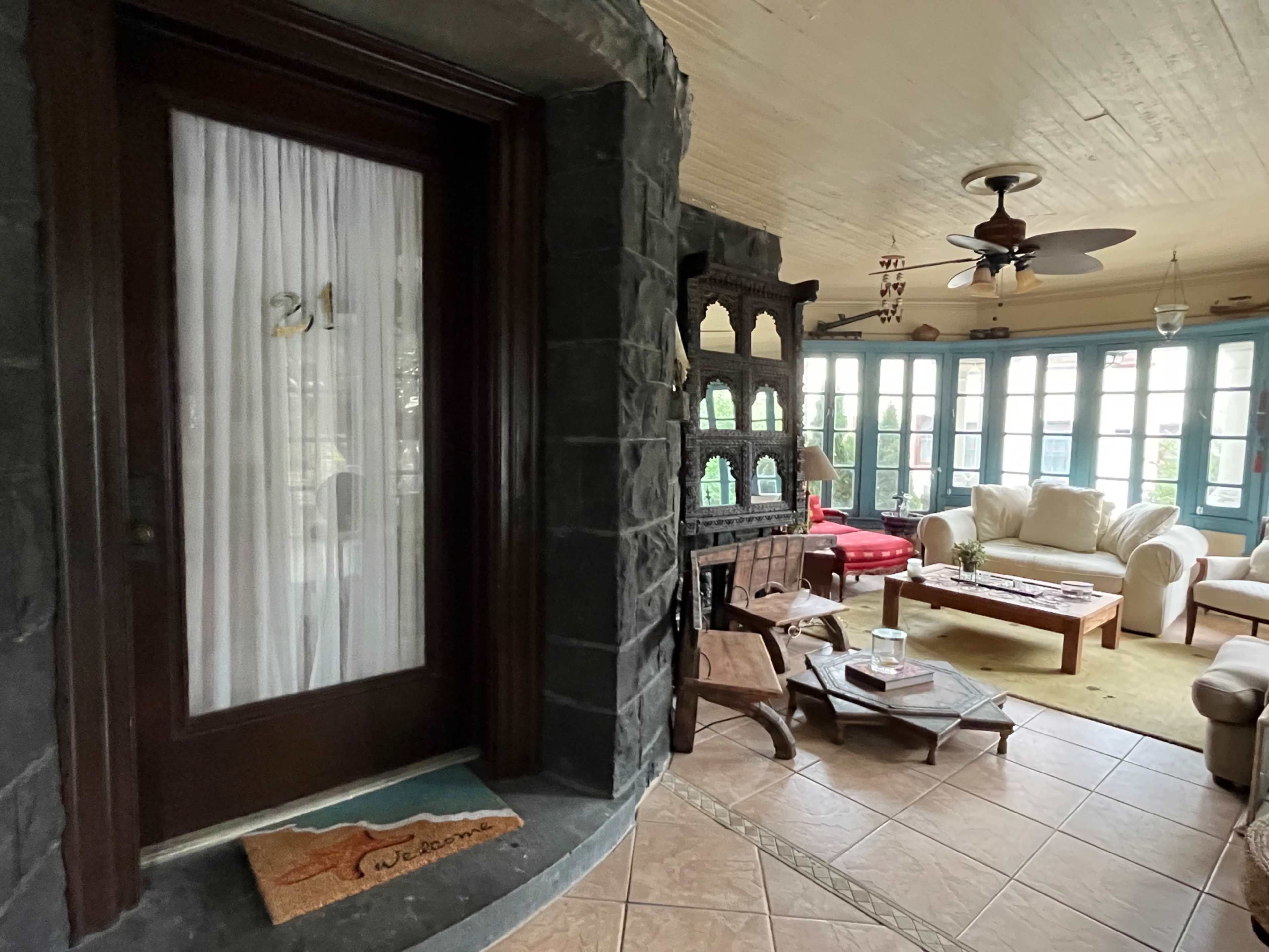 A cozy entryway features a front door with a decorative curtain and a welcome mat, leading into a bright room filled with light furniture and a large windowed area.
