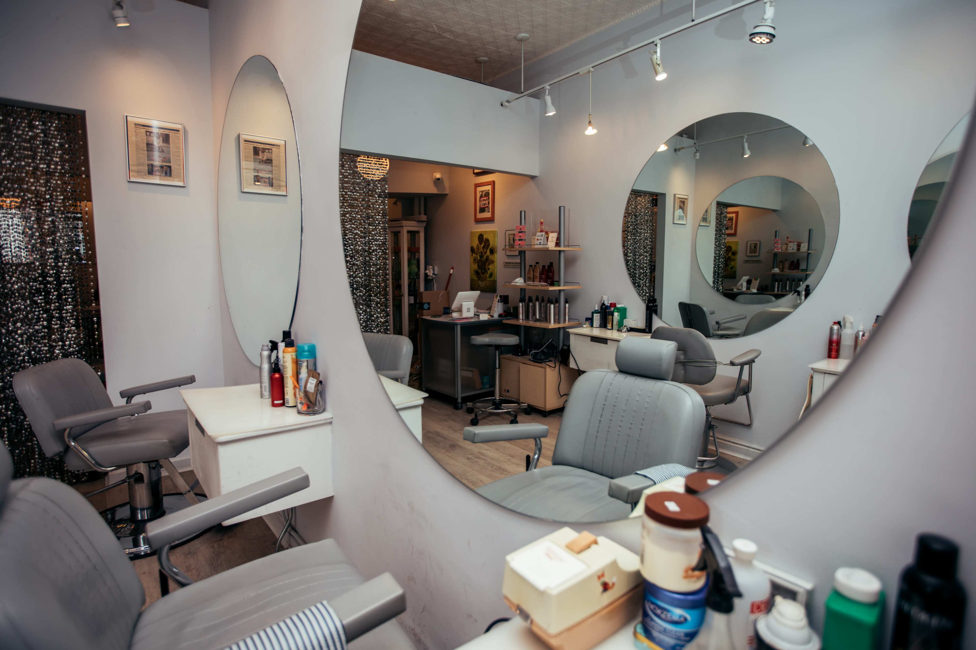 The image shows a modern hair salon with multiple styling stations reflected in large round mirrors and neatly arranged products on the counters.