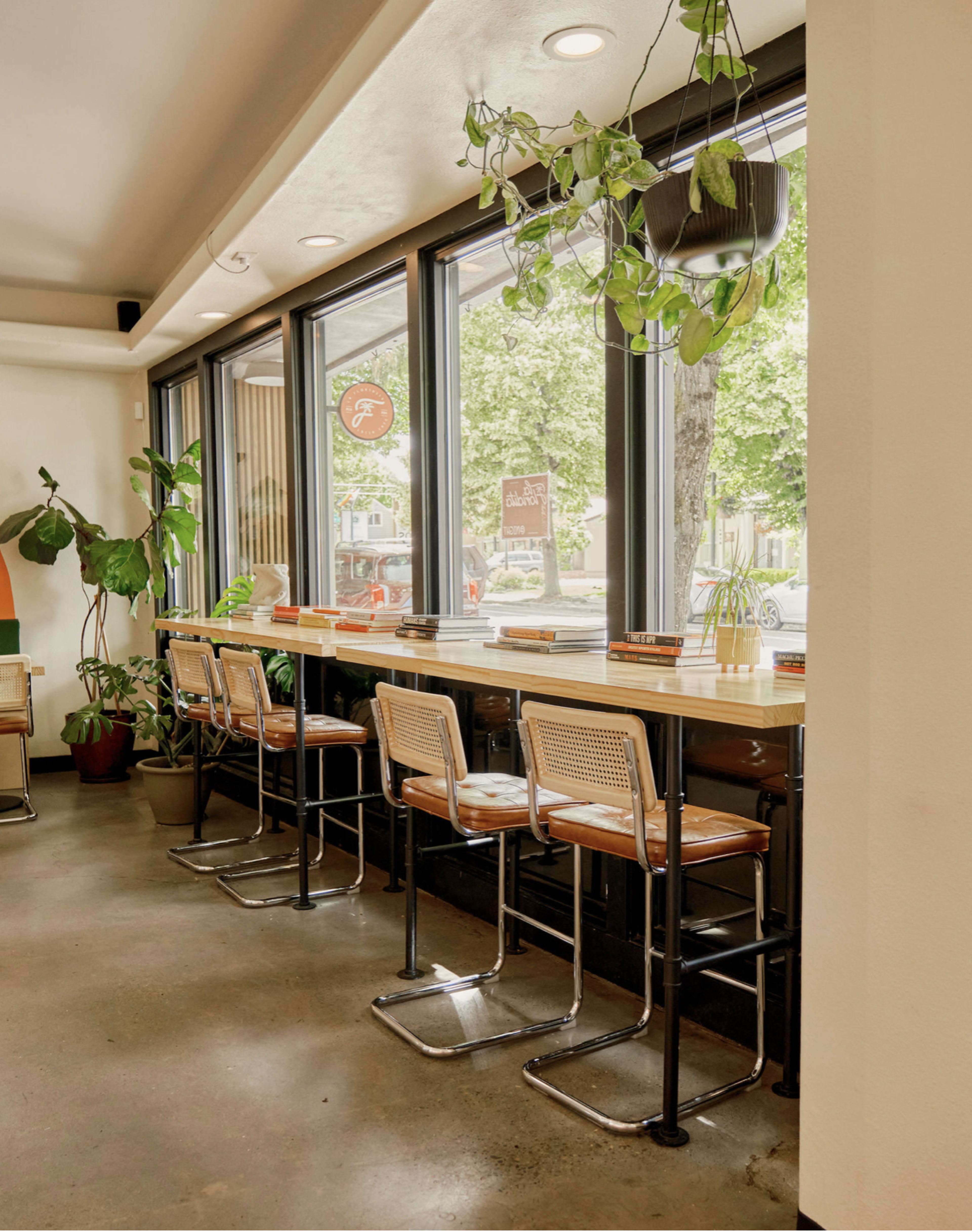 The image shows a bright café interior with a row of barstools facing large windows that overlook greenery outside.