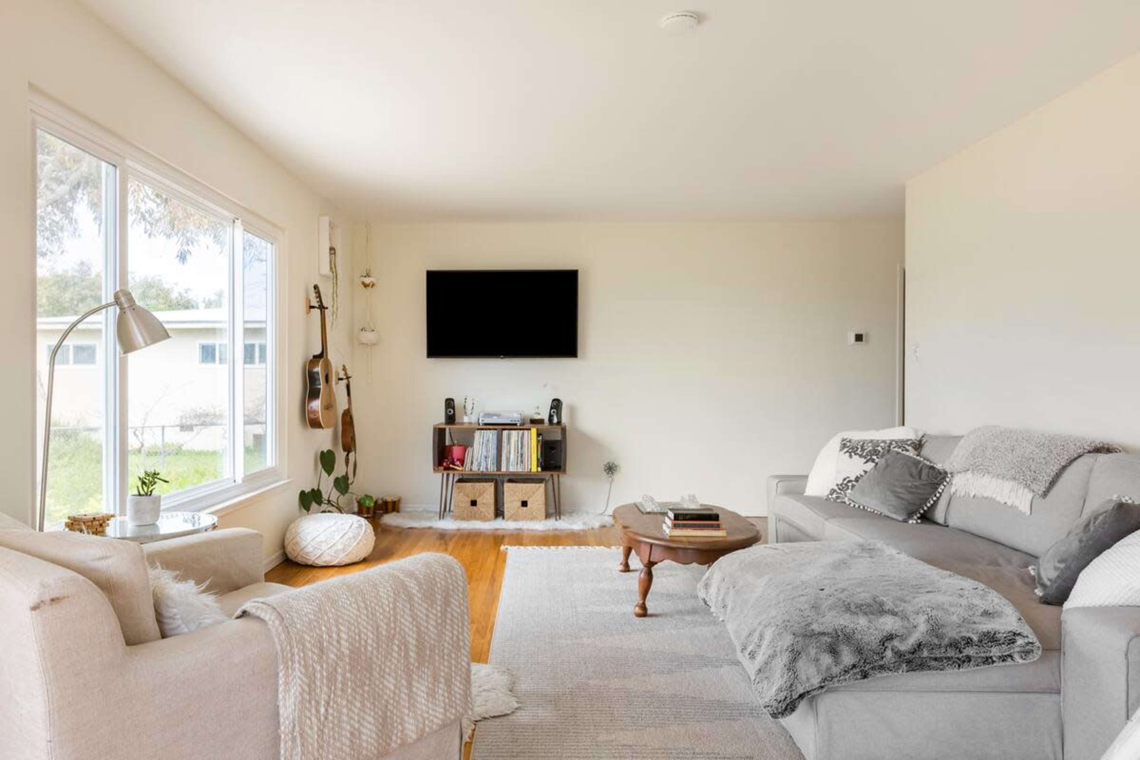 A light-filled living room features a gray sofa, a plush armchair, and a wooden coffee table, with a wall-mounted TV and shelves displaying books and decor.
