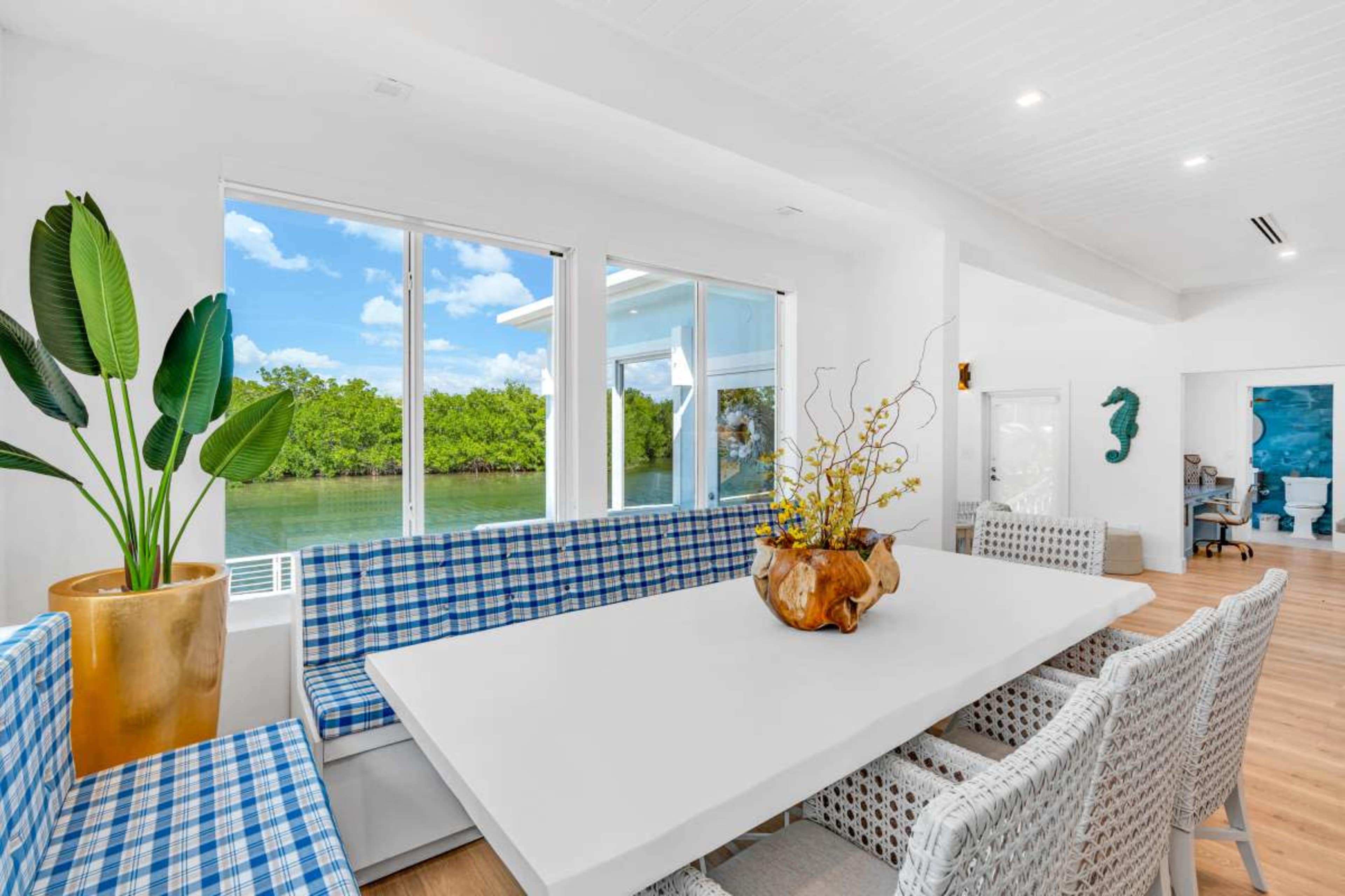A bright dining area features a large white table surrounded by woven chairs and a blue-checkered bench, with large windows overlooking a green landscape.