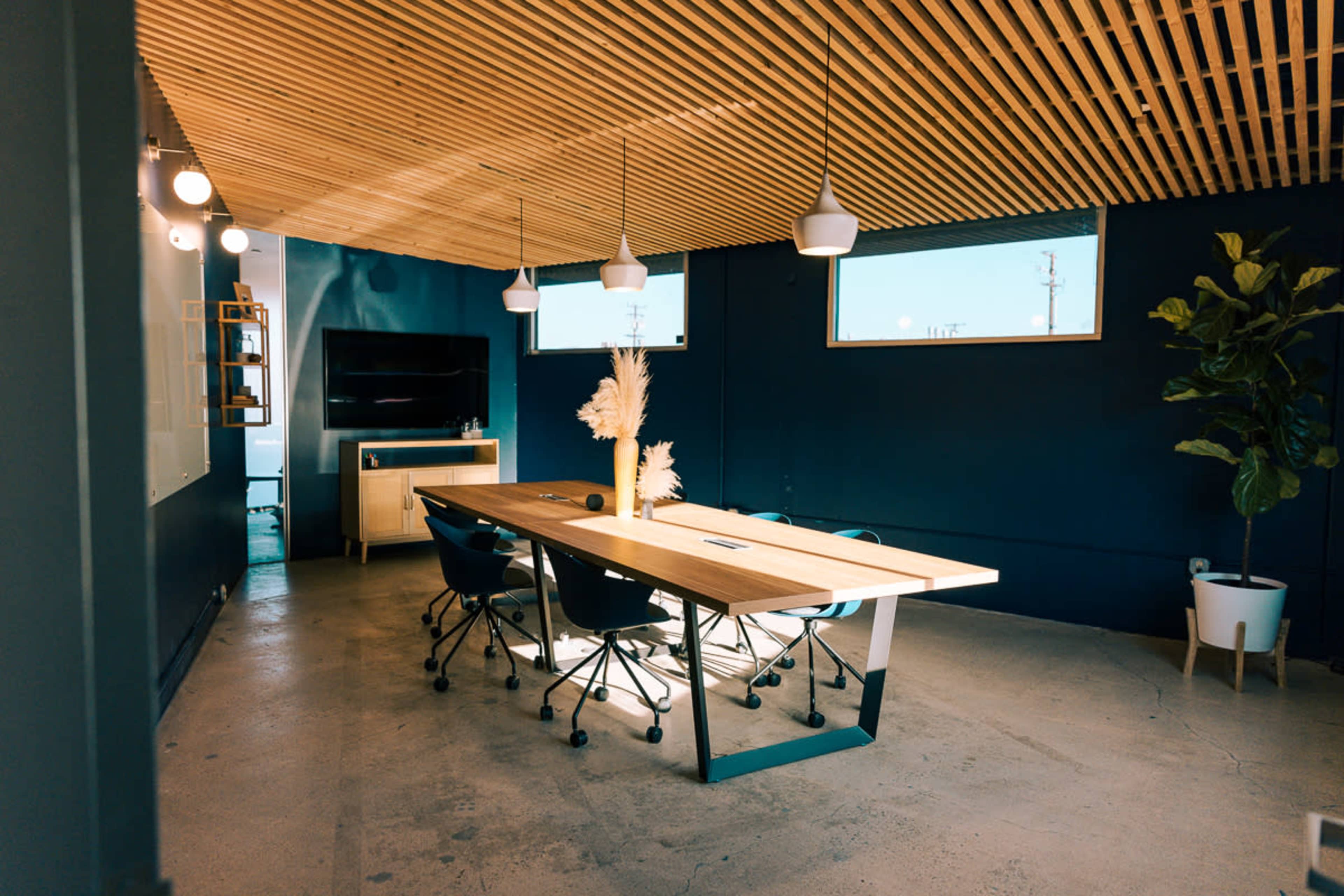 A modern conference room featuring a large wooden table surrounded by rolling chairs, with a plant and a wall-mounted TV in the background.